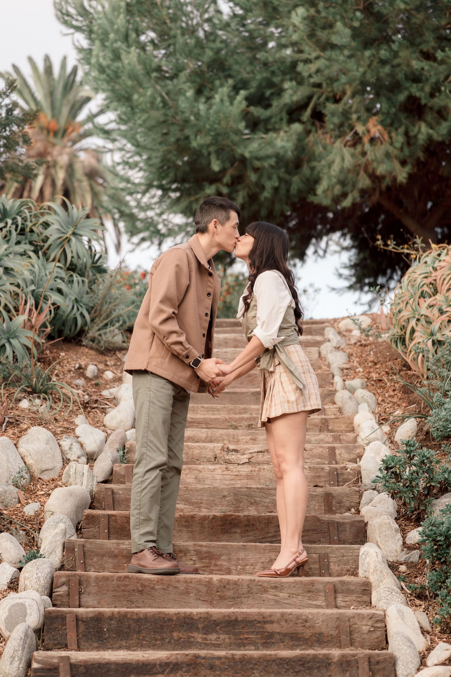 Couple shares a romantic kiss while holding hands on wooden steps surrounded by lush garden greenery.