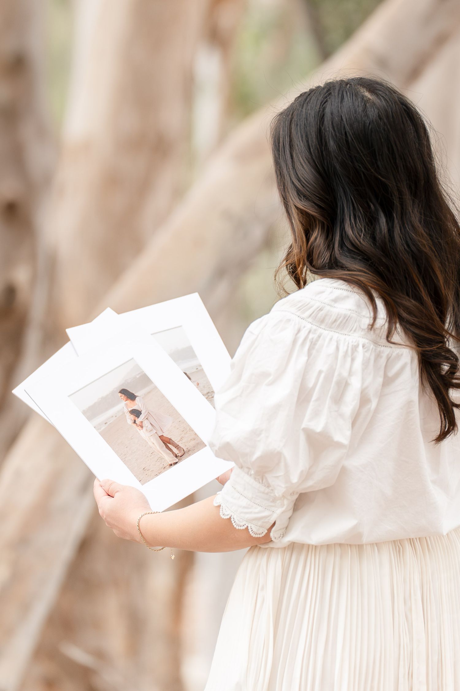 A young woman in a white blouse and skirt holds framed photos outdoors near a tree.