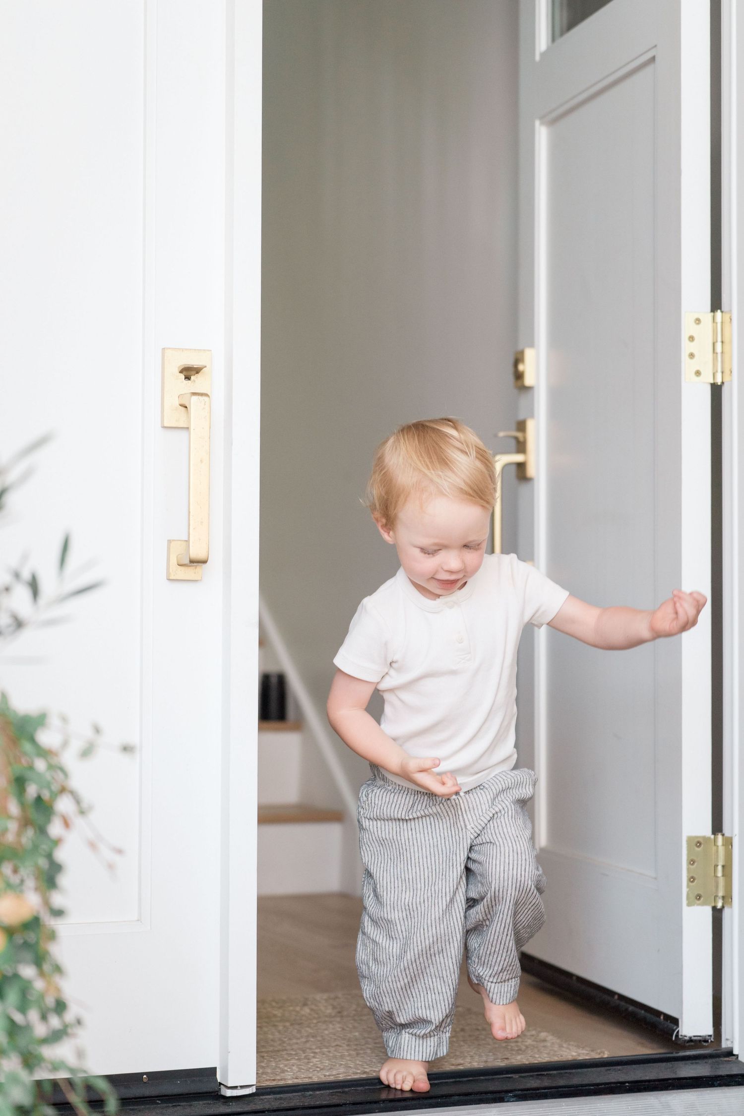 Blonde toddler standing barefoot in open white door with gold hardware.