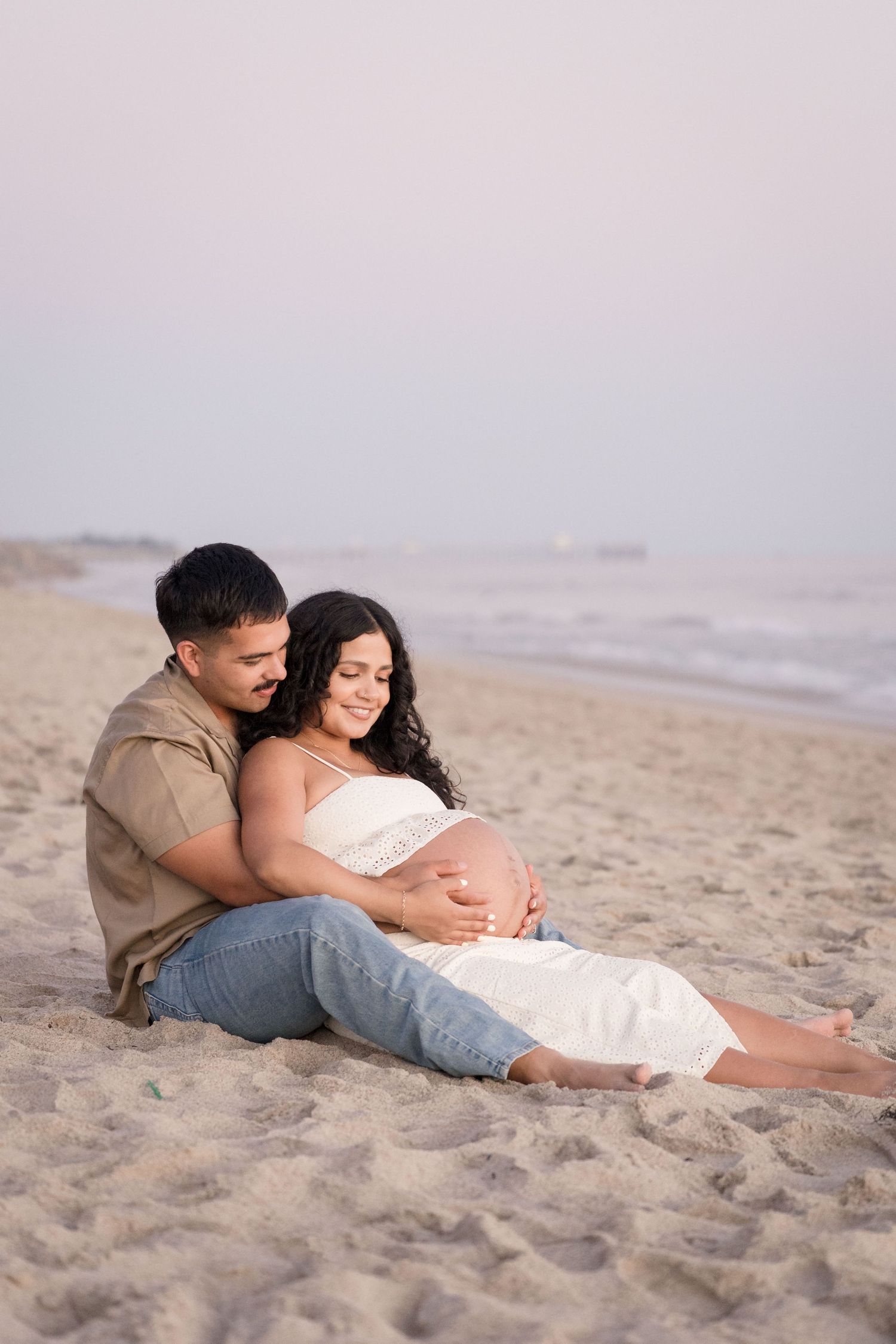 Expecting couple sitting on beach at dusk, man embracing pregnant woman from behind.
