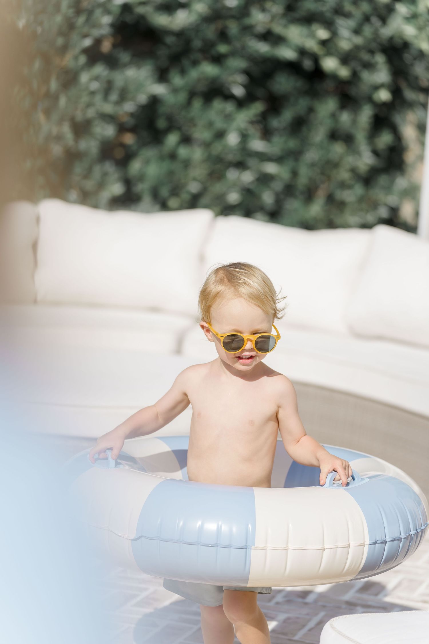Cute blonde toddler wearing yellow sunglasses stands inside inflatable pool ring outdoors.