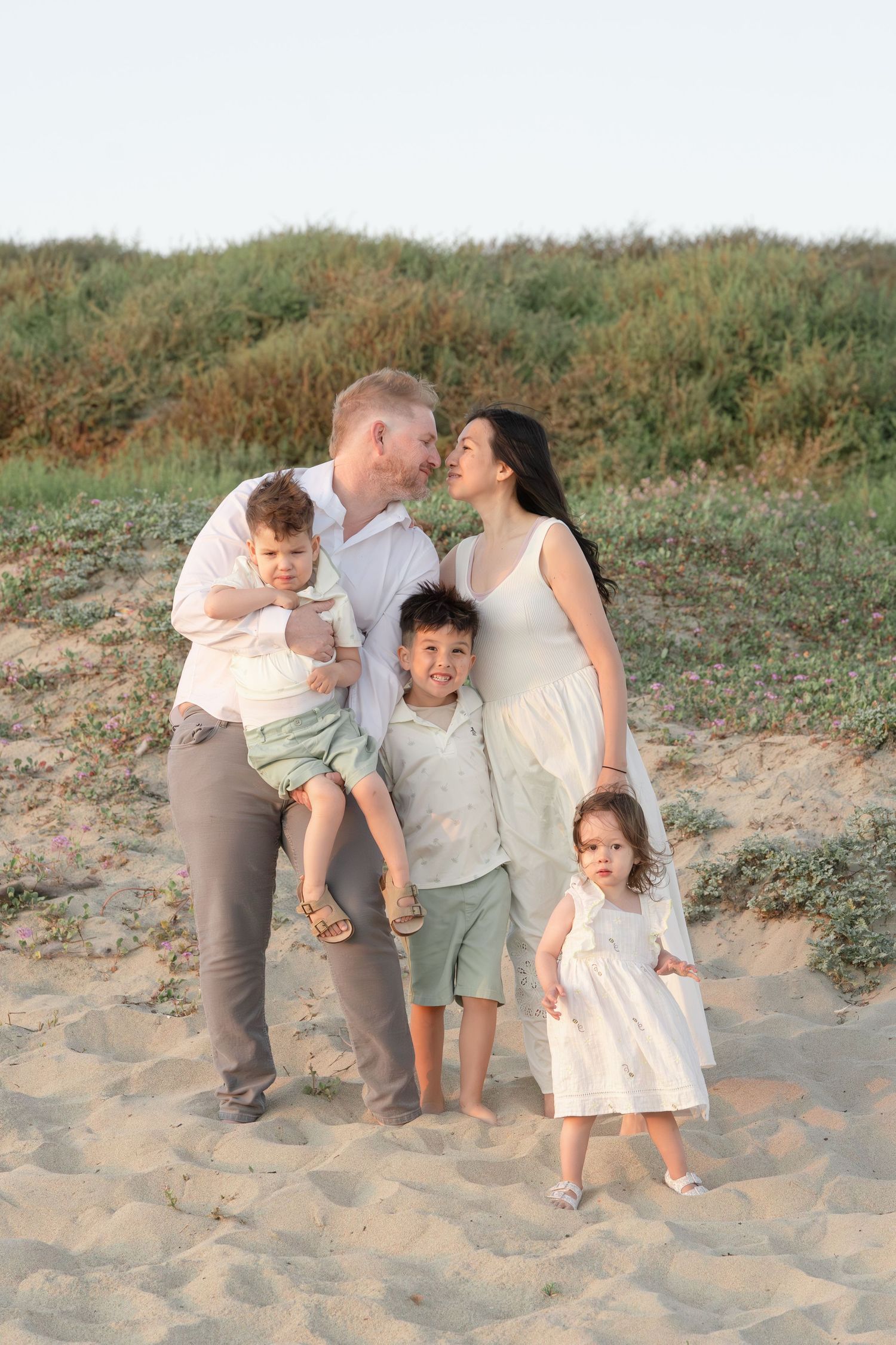 Interracial family of five on sandy beach dunes, parents kissing with three young children.
