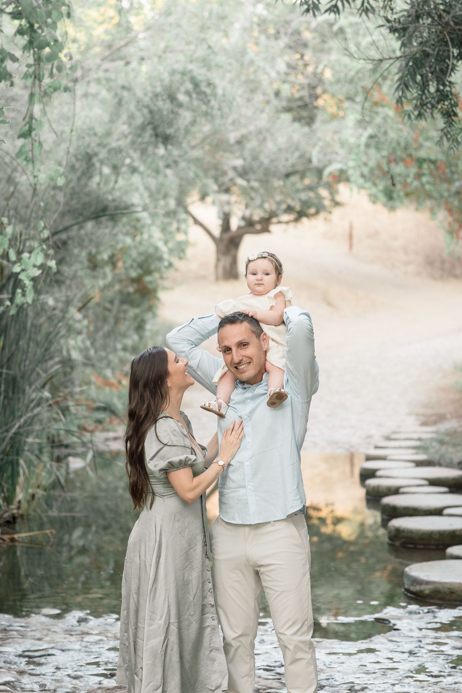 Happy family with baby outdoors near stepping stone pond, parents in neutral tones holding infant.
