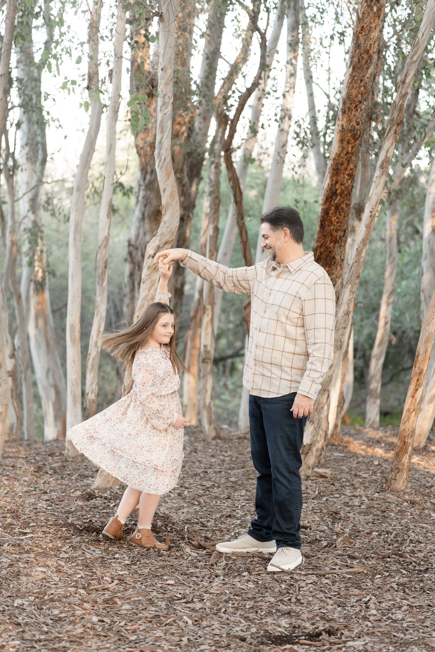A father twirls his young daughter in a floral dress among eucalyptus trees in a warm outdoor setting.