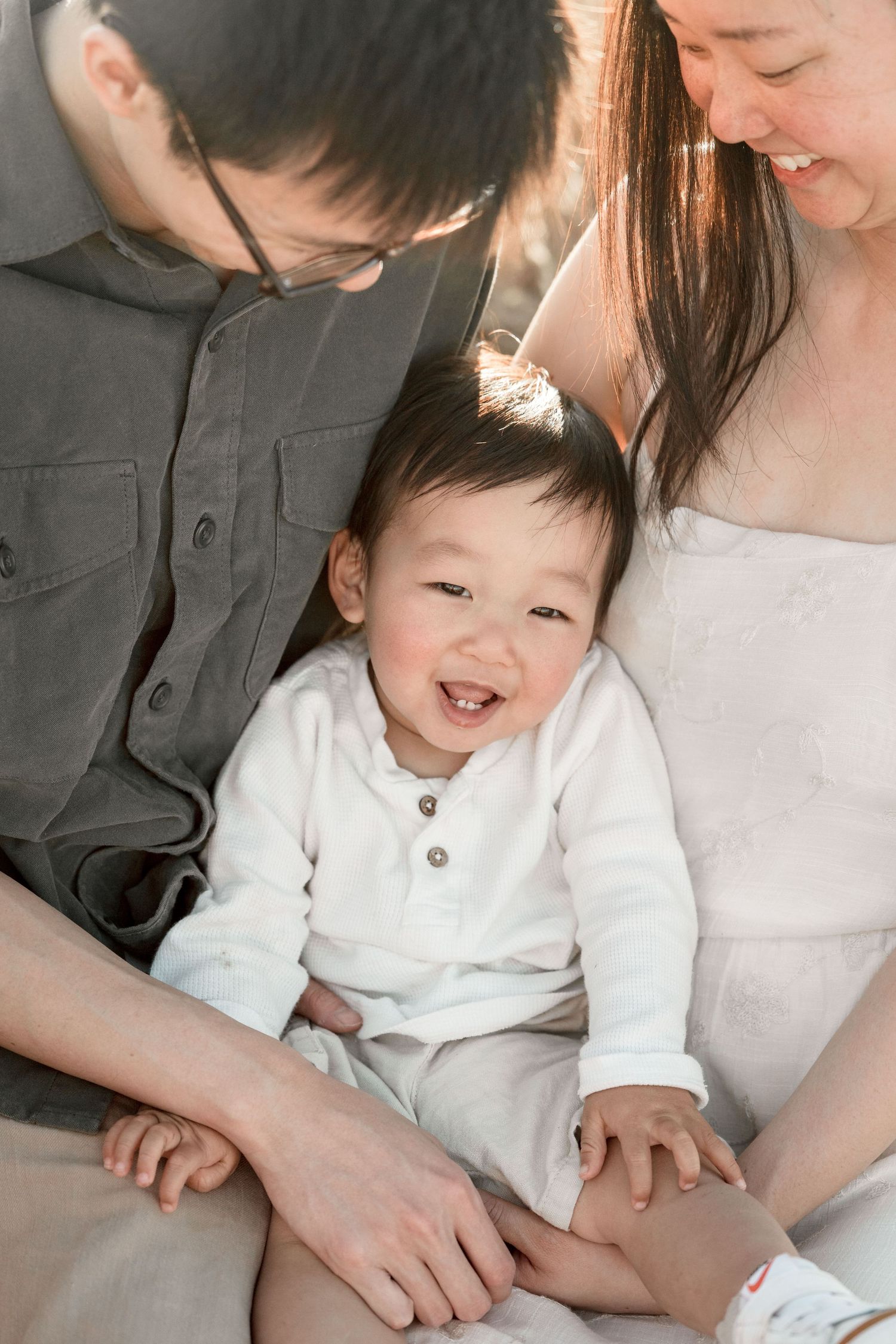 A smiling Asian baby in white sits between his parents, laughing joyfully in warm sunlight.