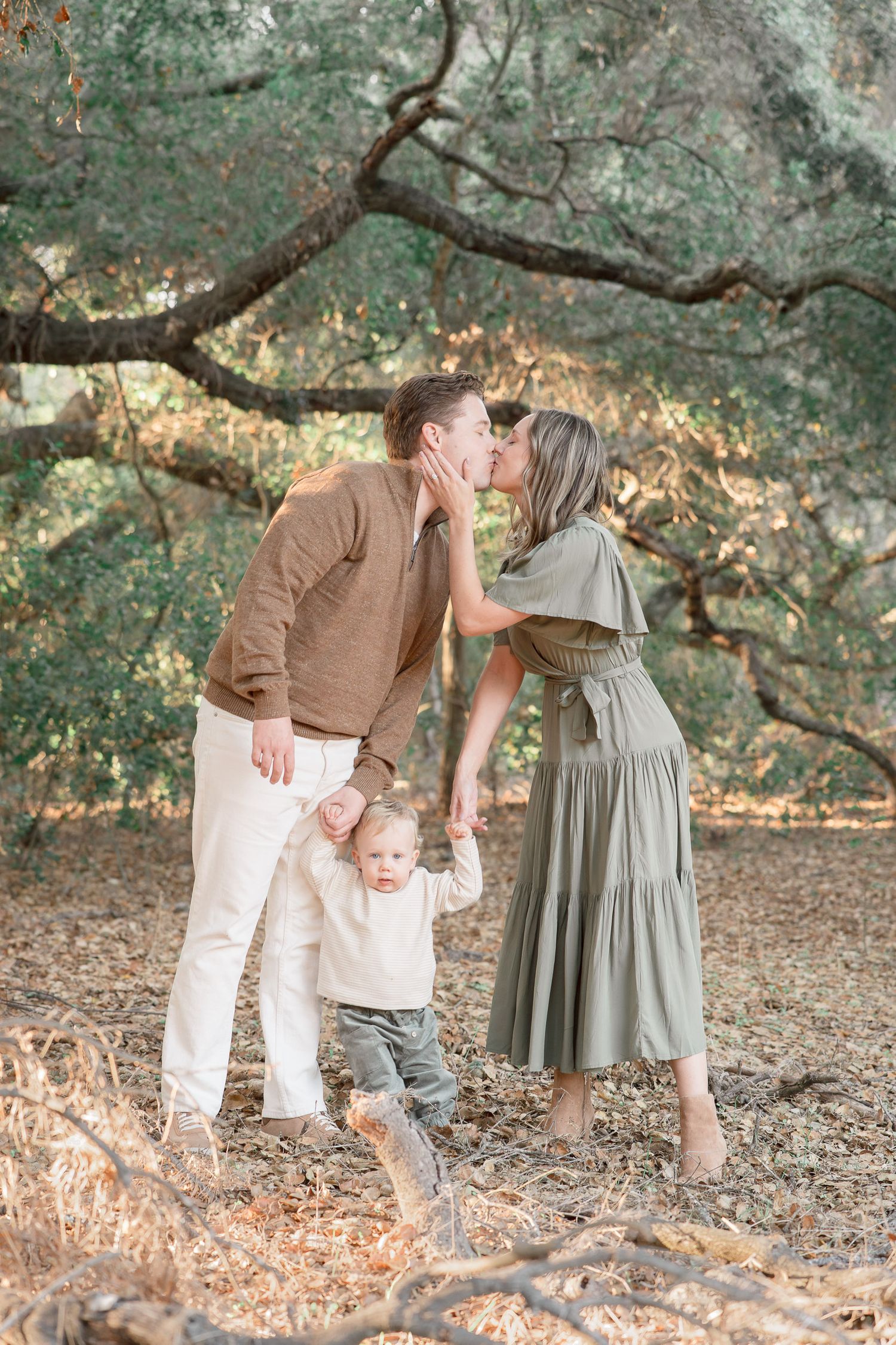 A couple kisses while holding their toddler's hands beneath sprawling oak trees in a warm, natural outdoor setting.