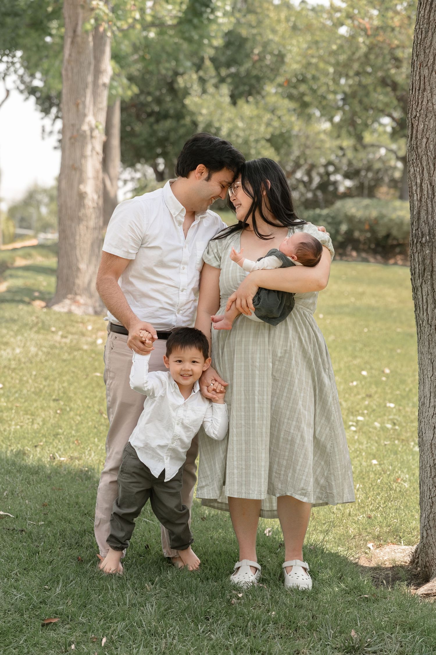 Happy Asian family with toddler son and newborn baby posing outdoors in park under green trees.