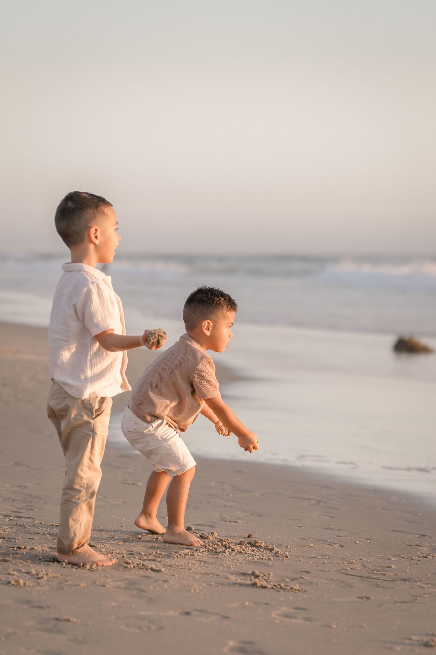 Two young boys standing on a beach at sunset, one holding a rock while the other prepares to throw.