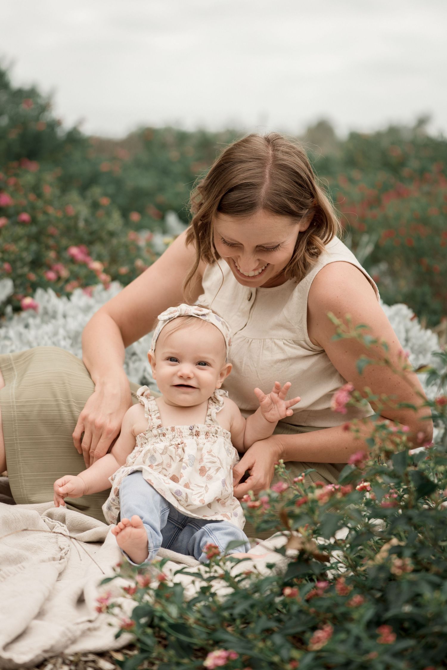 A smiling mother sits in a flower field with her happy baby girl wearing a floral outfit and white headband.
