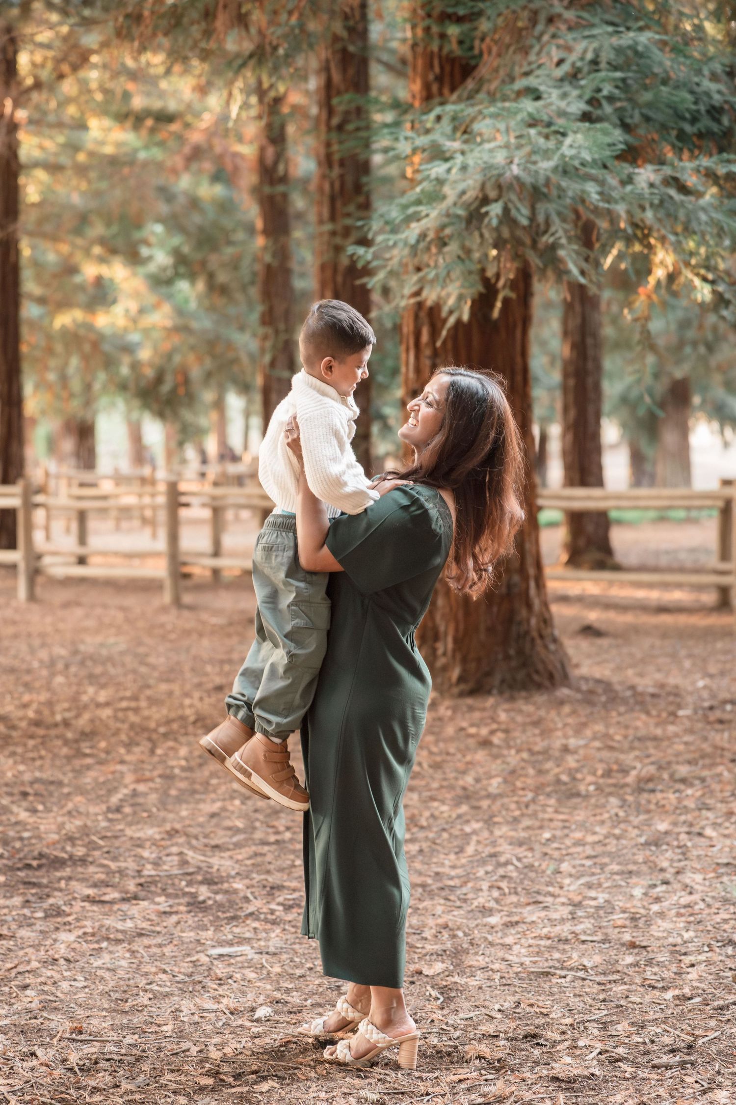 A smiling mother in a green dress lifts her young son in a white sweater among tall redwood trees.