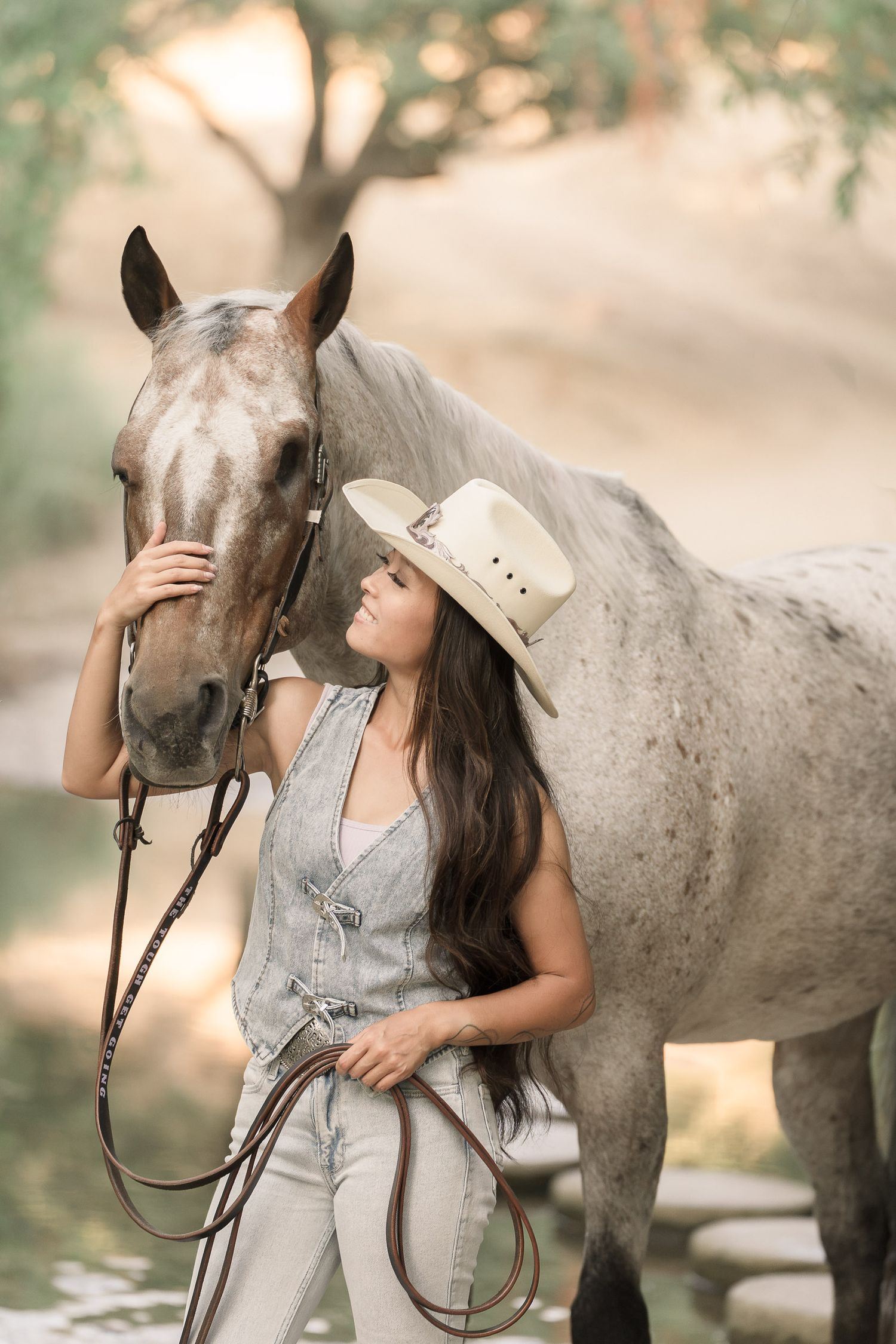 A woman in a cowboy hat and denim outfit tenderly holds a gray horse by the reins outdoors.