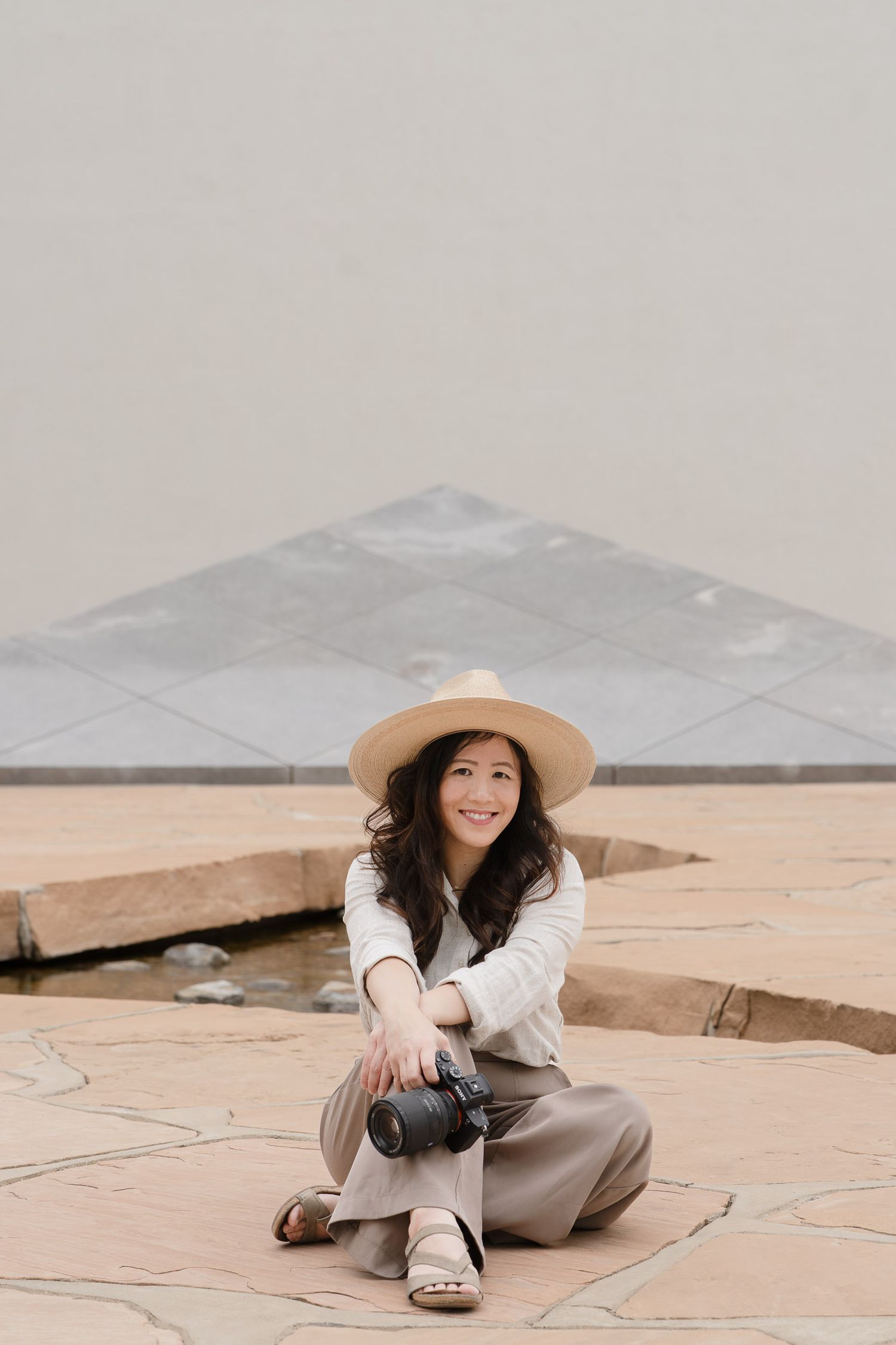 Young woman in a straw hat sits cross-legged on stone pavement holding a camera, smiling outdoors.