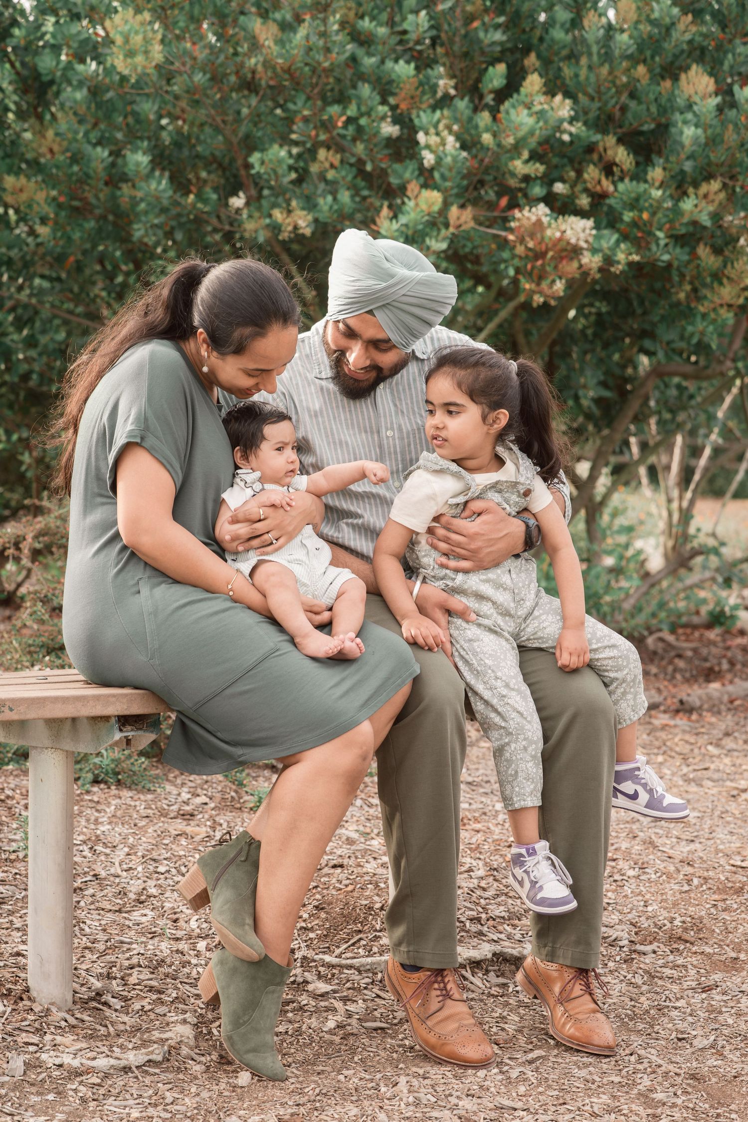 South Asian family of four with baby and toddler sitting together outdoors near green trees.