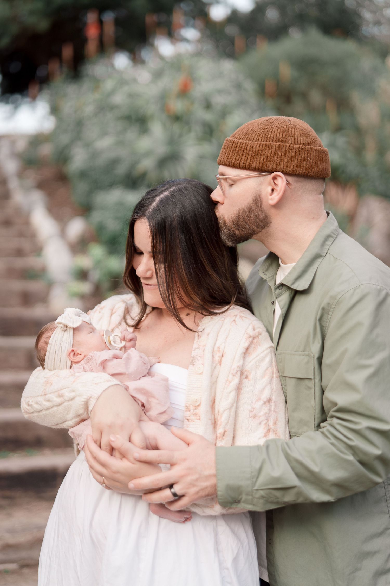 Father in green jacket and brown beanie kisses mother holding newborn baby outdoors near stone steps.