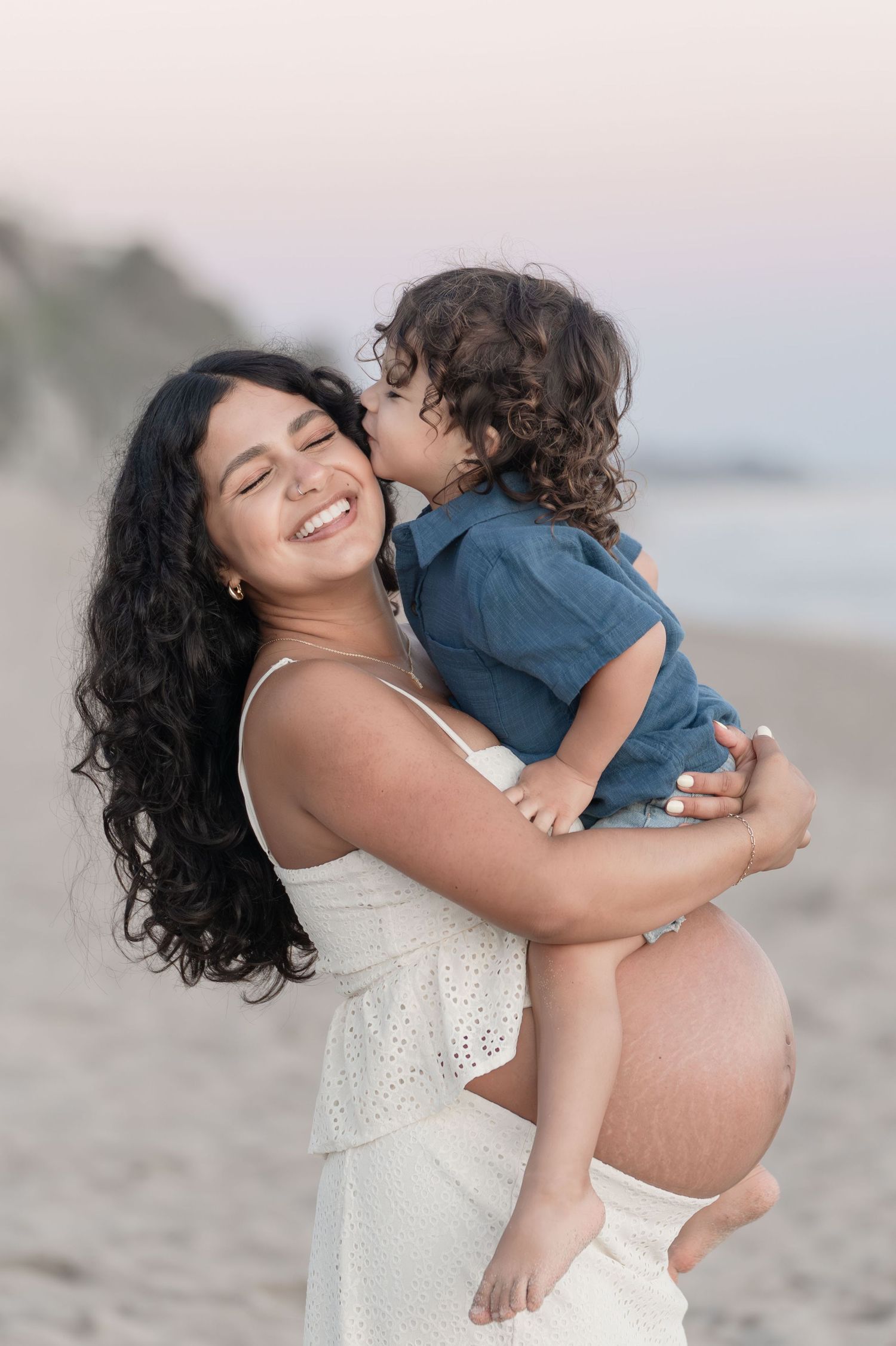 Smiling pregnant woman in white dress holding curly-haired toddler who kisses her cheek on sandy beach.