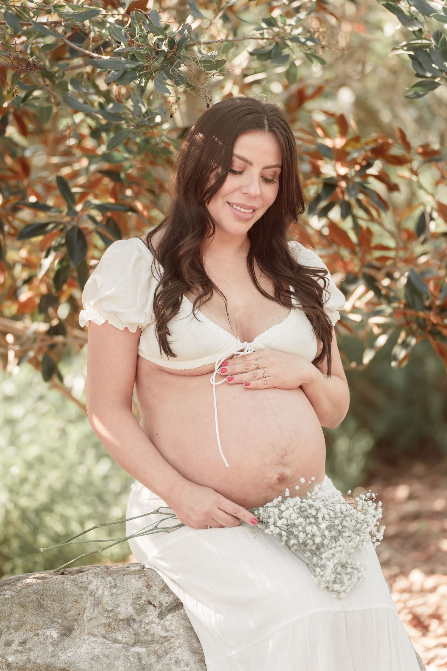 Pregnant woman in white dress holding baby's breath flowers, cradling bare belly outdoors among lush green foliage.