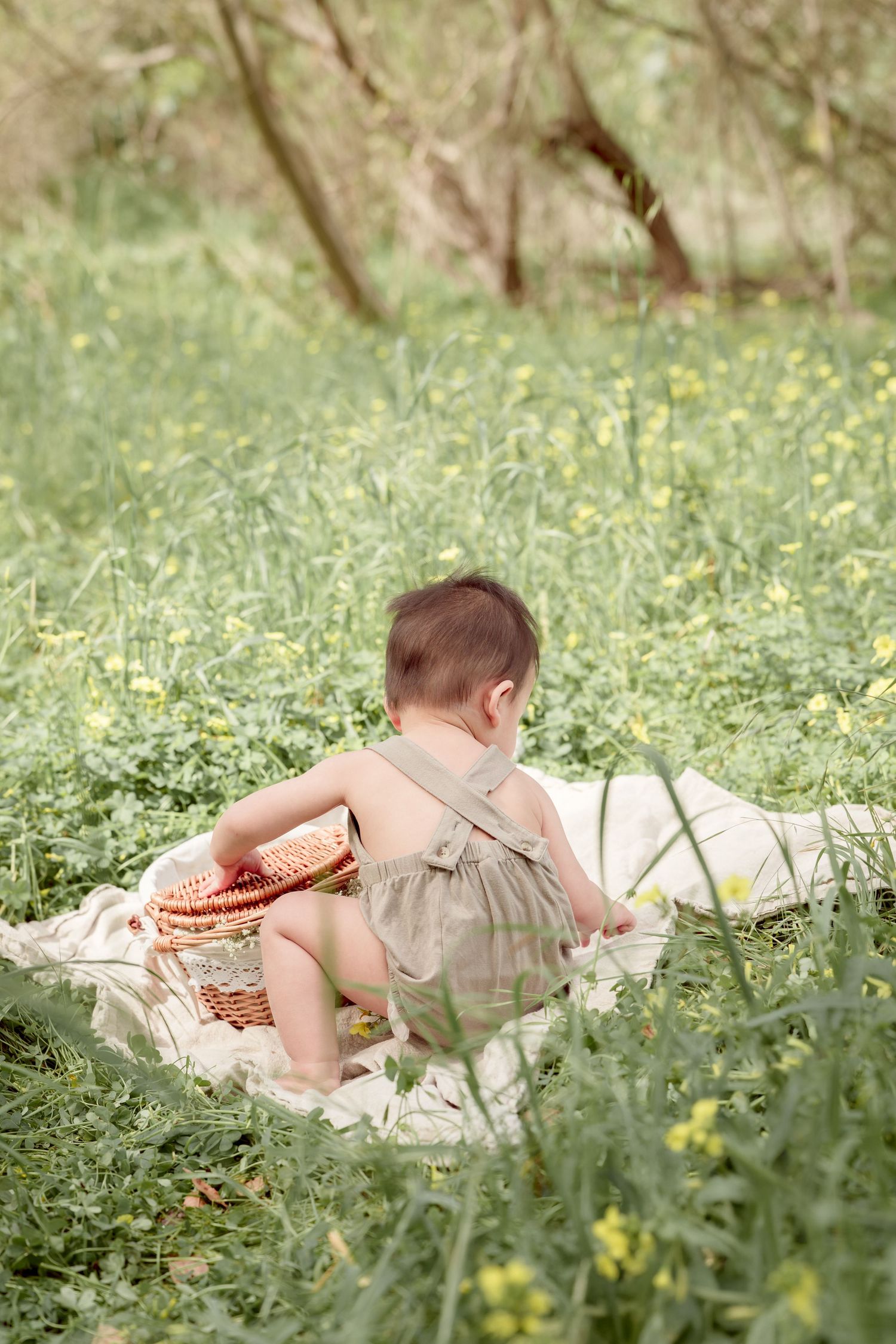 A baby in a green romper sits with their back to the camera next to a wicker basket in a lush green meadow.