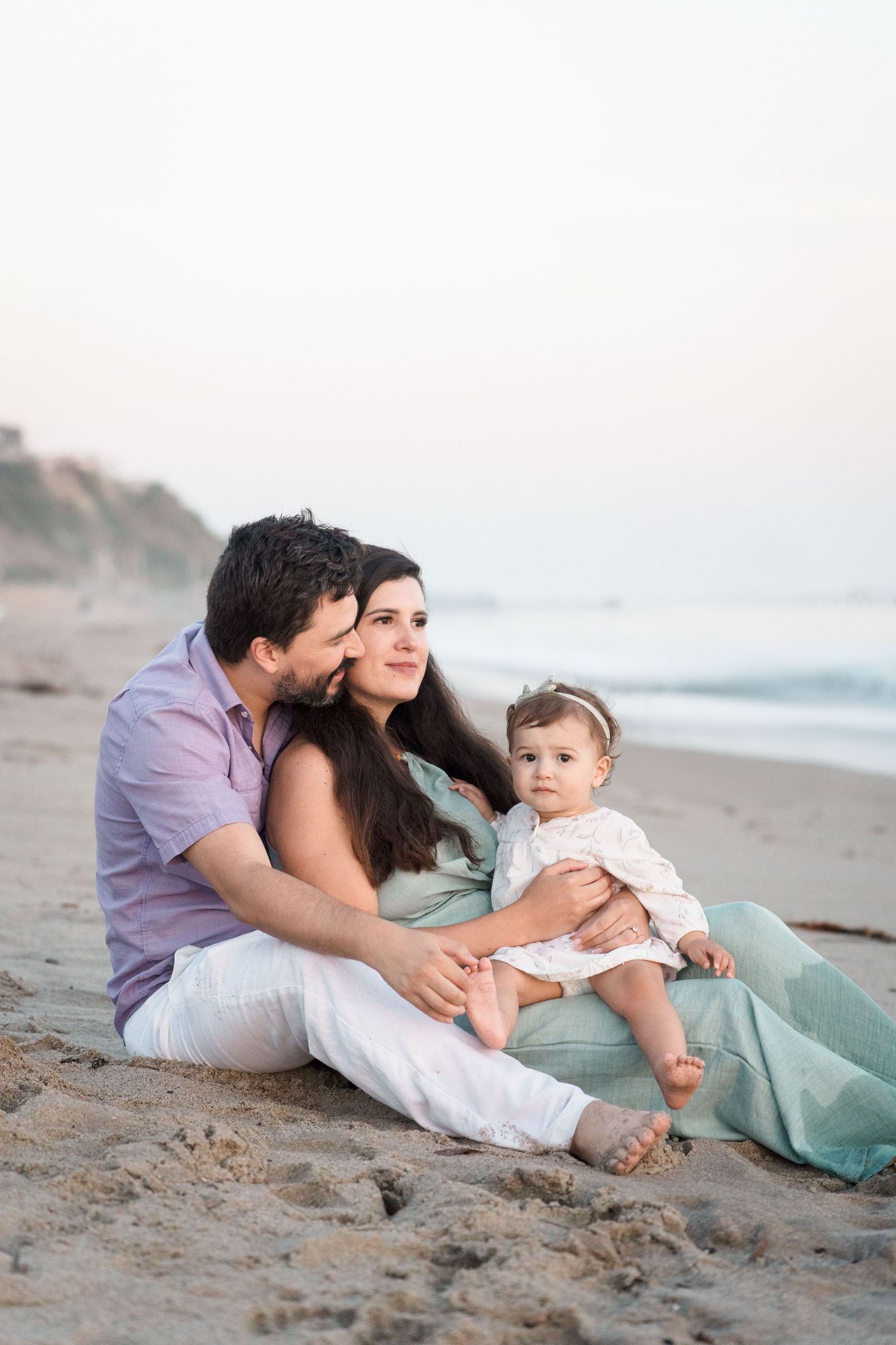 Family of three sitting on sandy beach at sunset, dad kissing mom while holding baby girl.