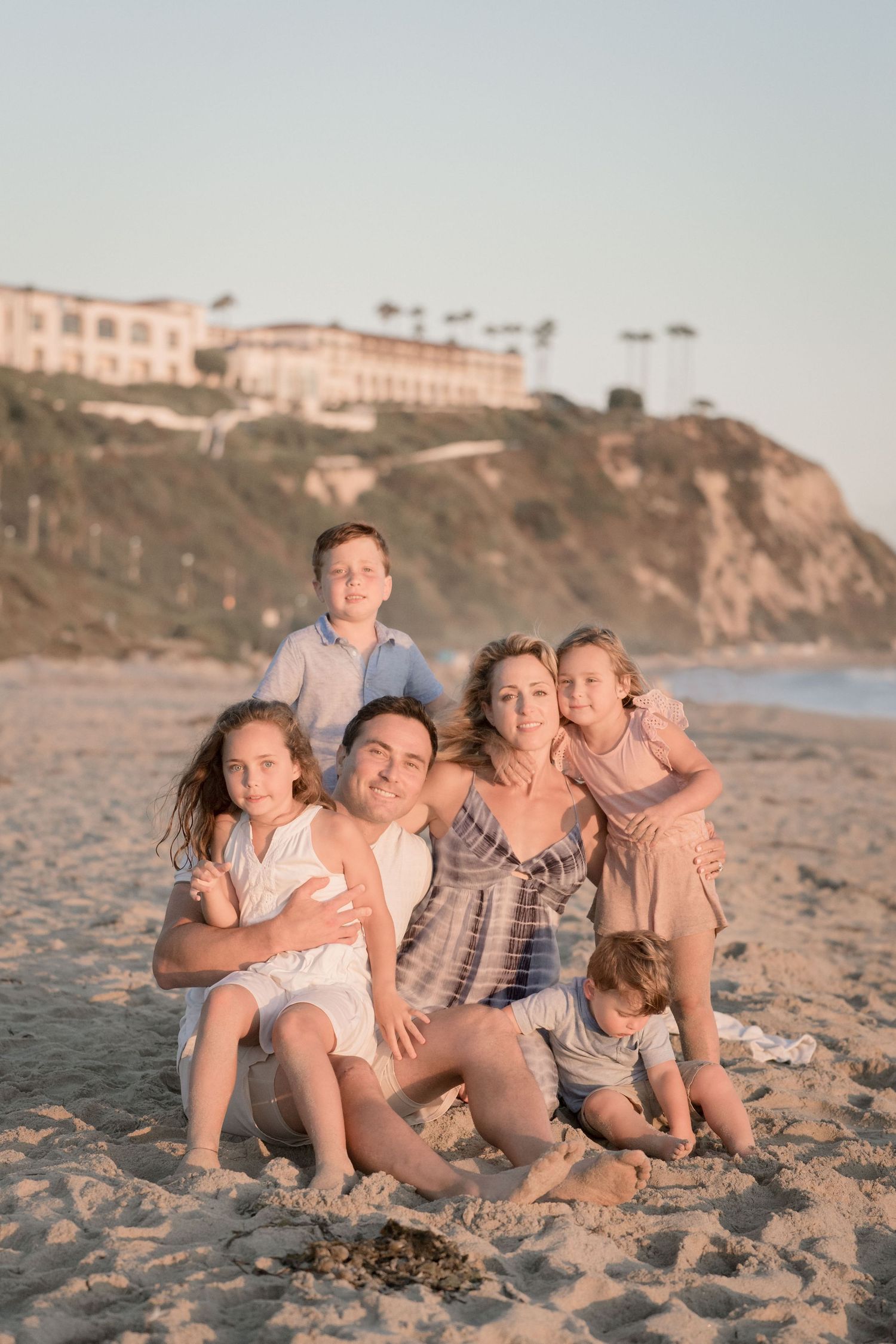 Family of six sitting together on a sandy beach at golden hour with coastal cliffs behind them.