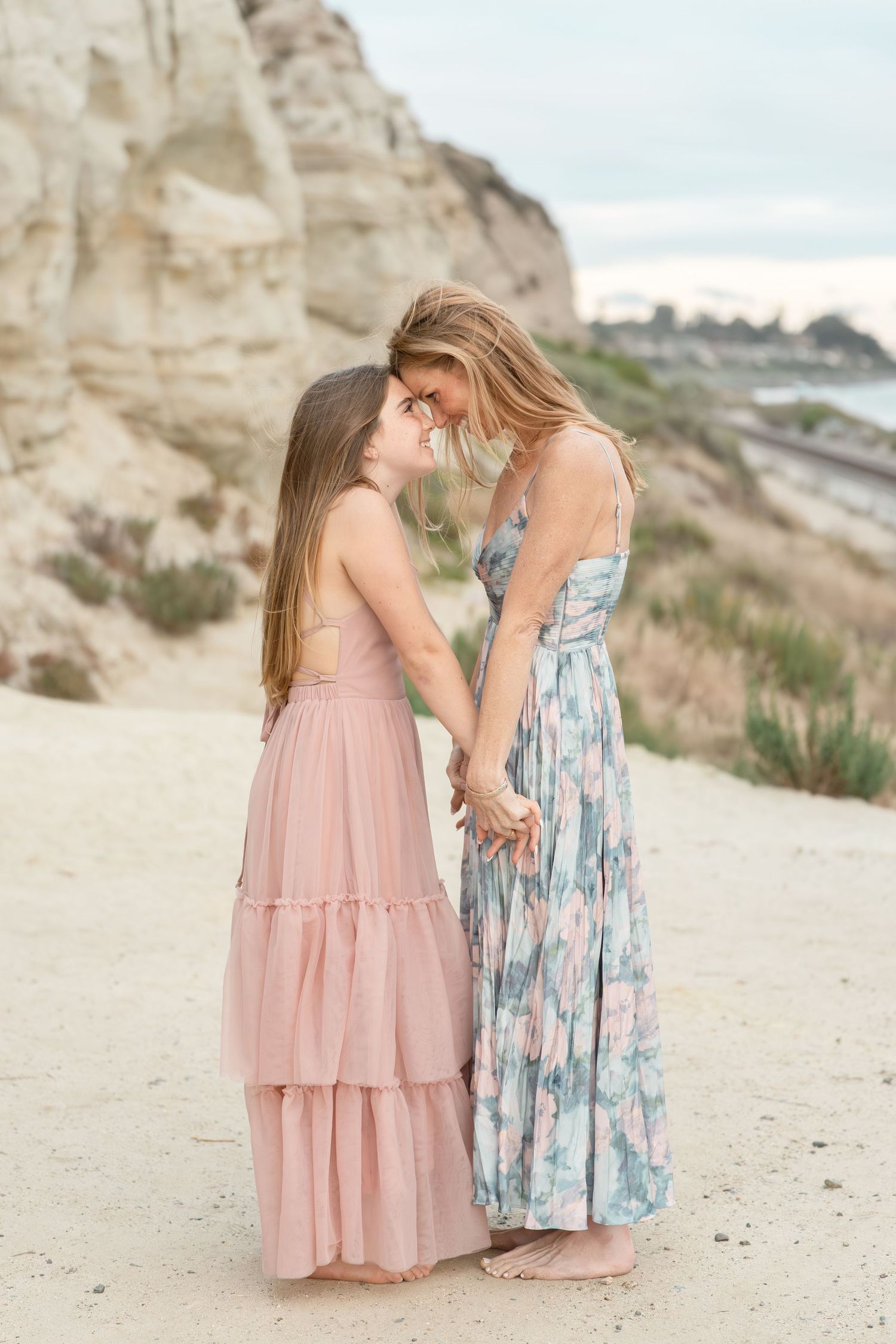 A mother and daughter in matching maxi dresses hold hands and touch foreheads on a sandy beach with cliffs behind them.