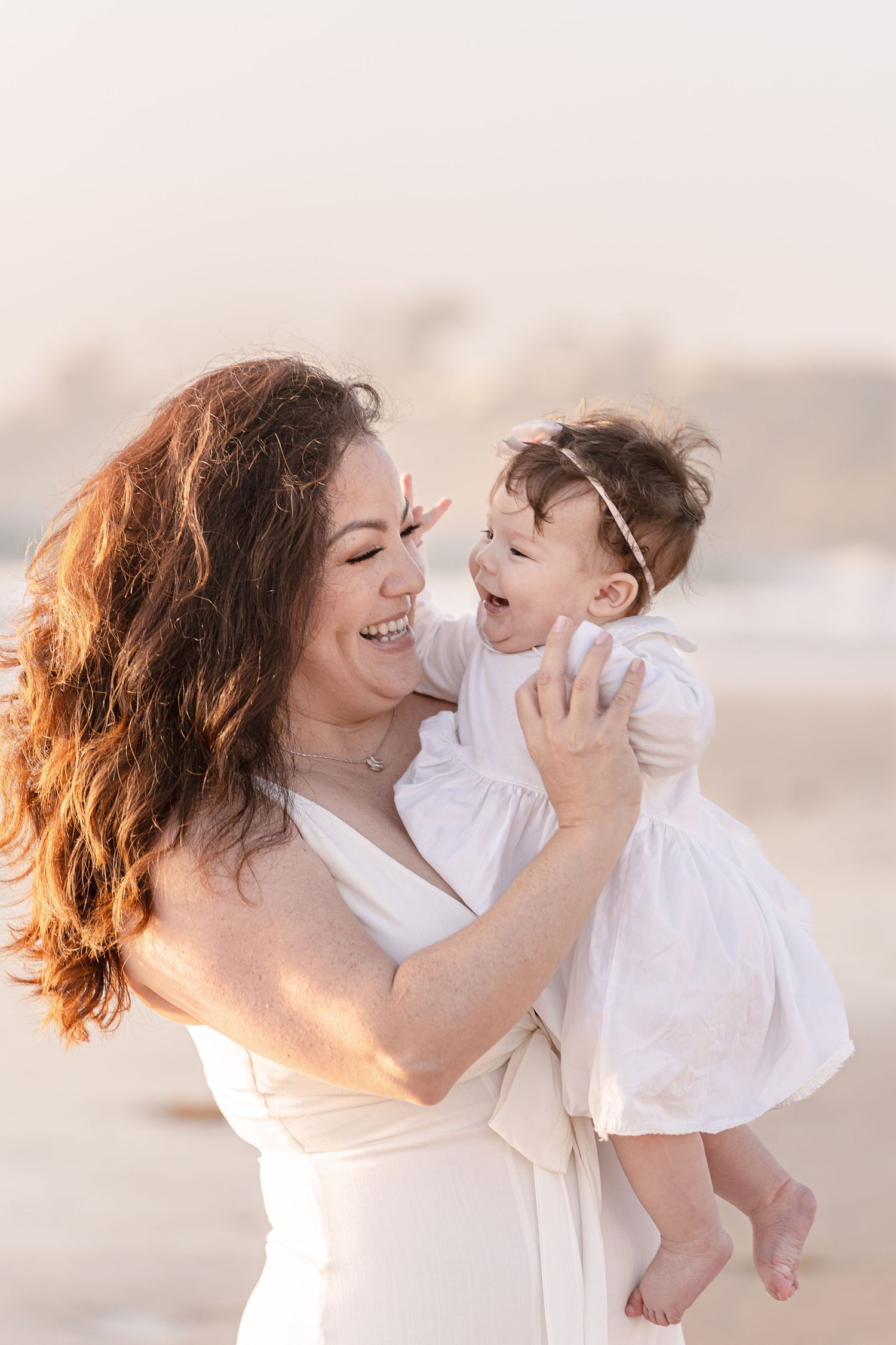 Laughing mother with curly red hair holds giggling baby girl on a sunny beach.