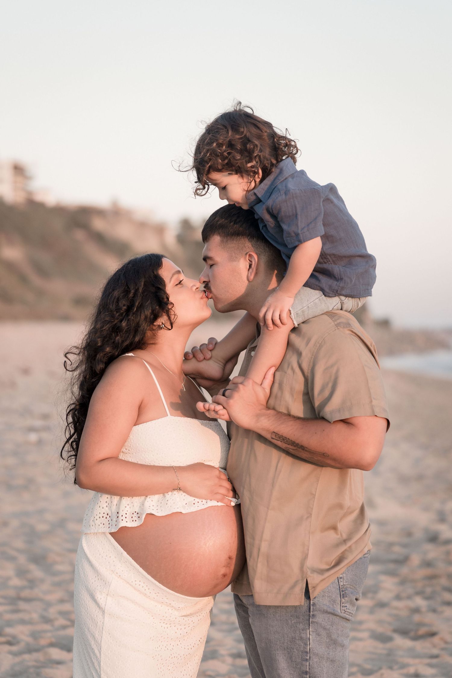 Pregnant woman in white dress kissing toddler on father's shoulders at sunset beach.