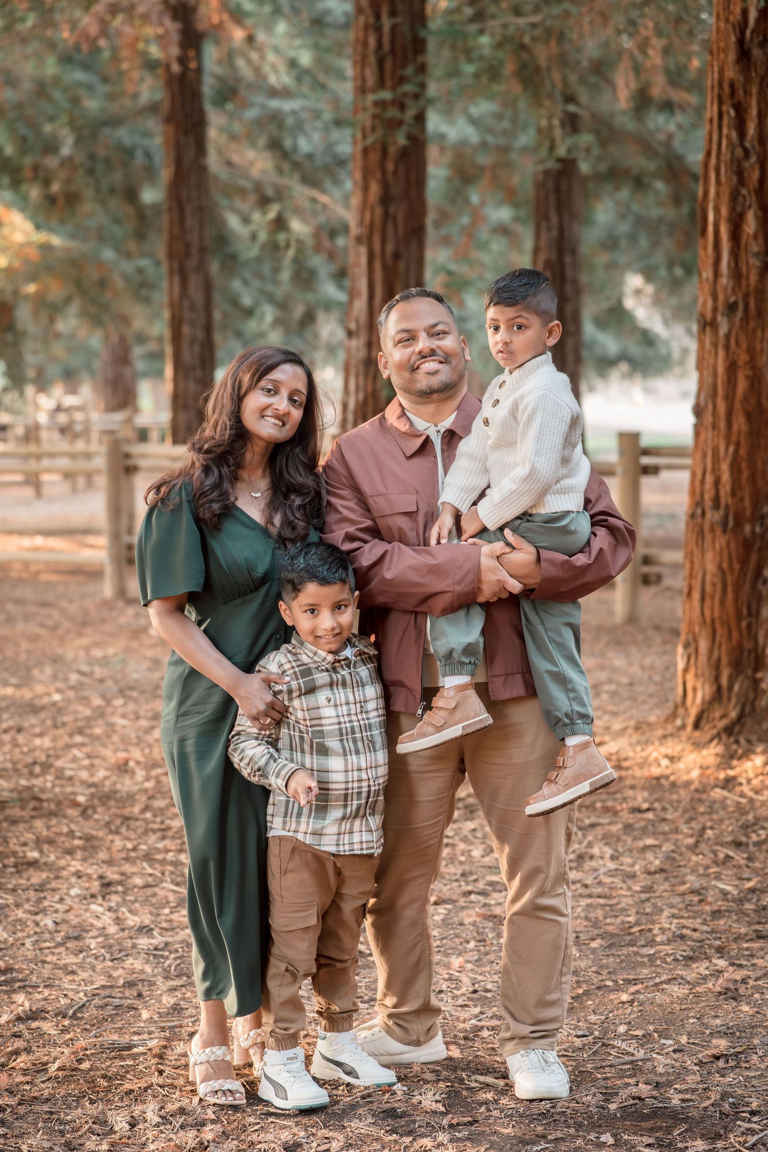 A happy South Asian family of four poses together in a redwood forest, parents holding their two young sons.