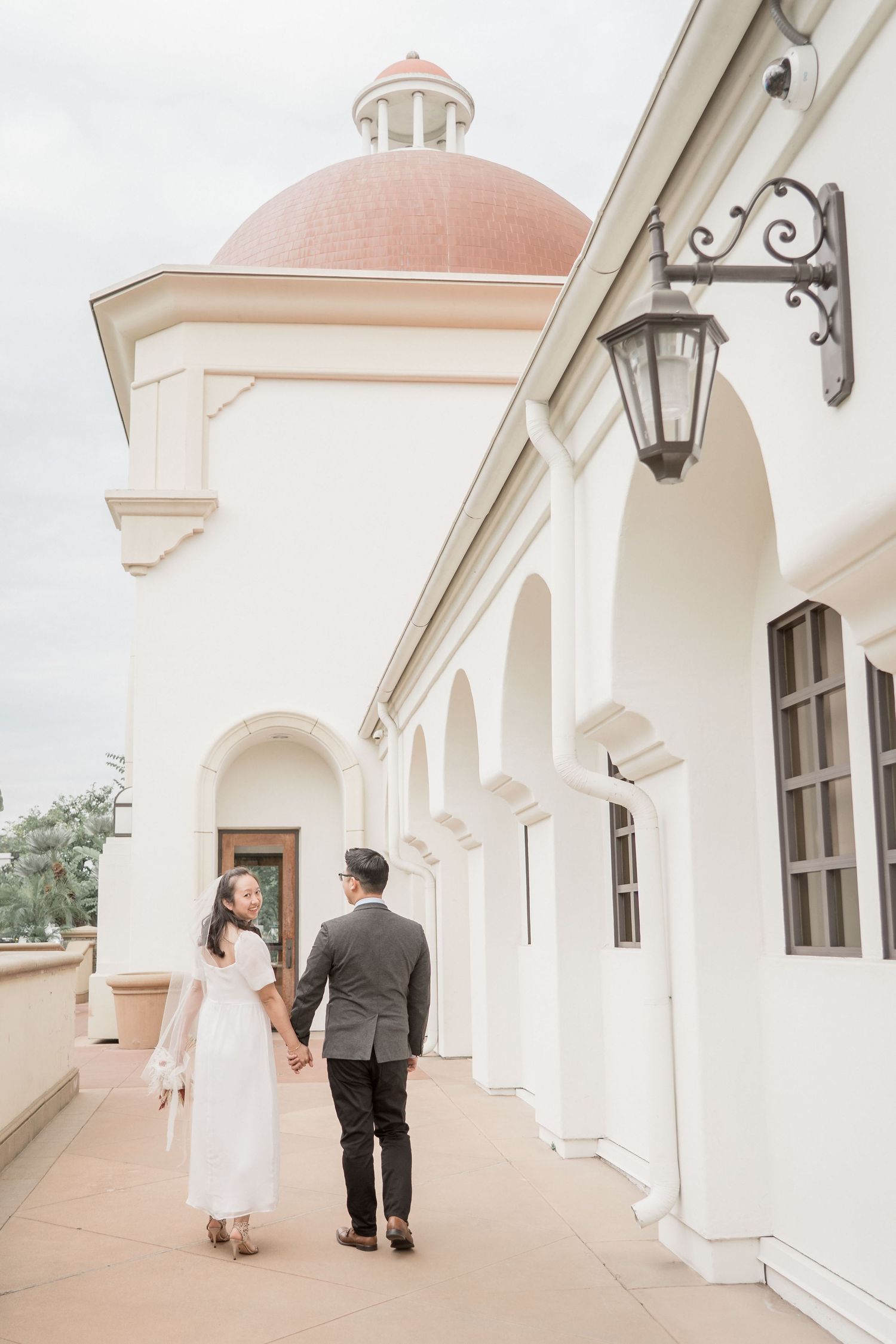 Newlywed couple holding hands walking along arched corridor of white building with red dome roof.