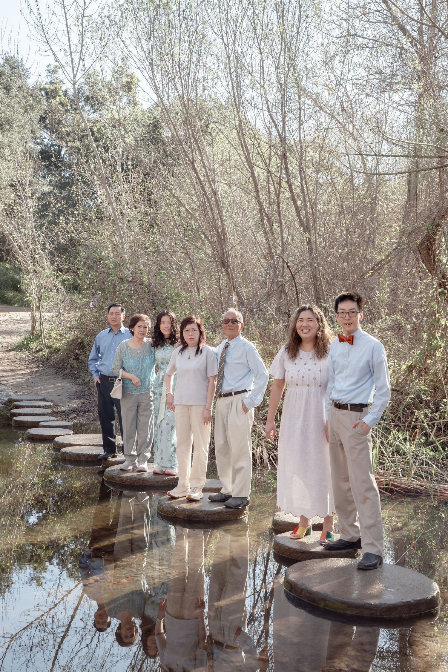 Multi-generational Asian family standing on stepping stones over a reflective stream in a winter park.