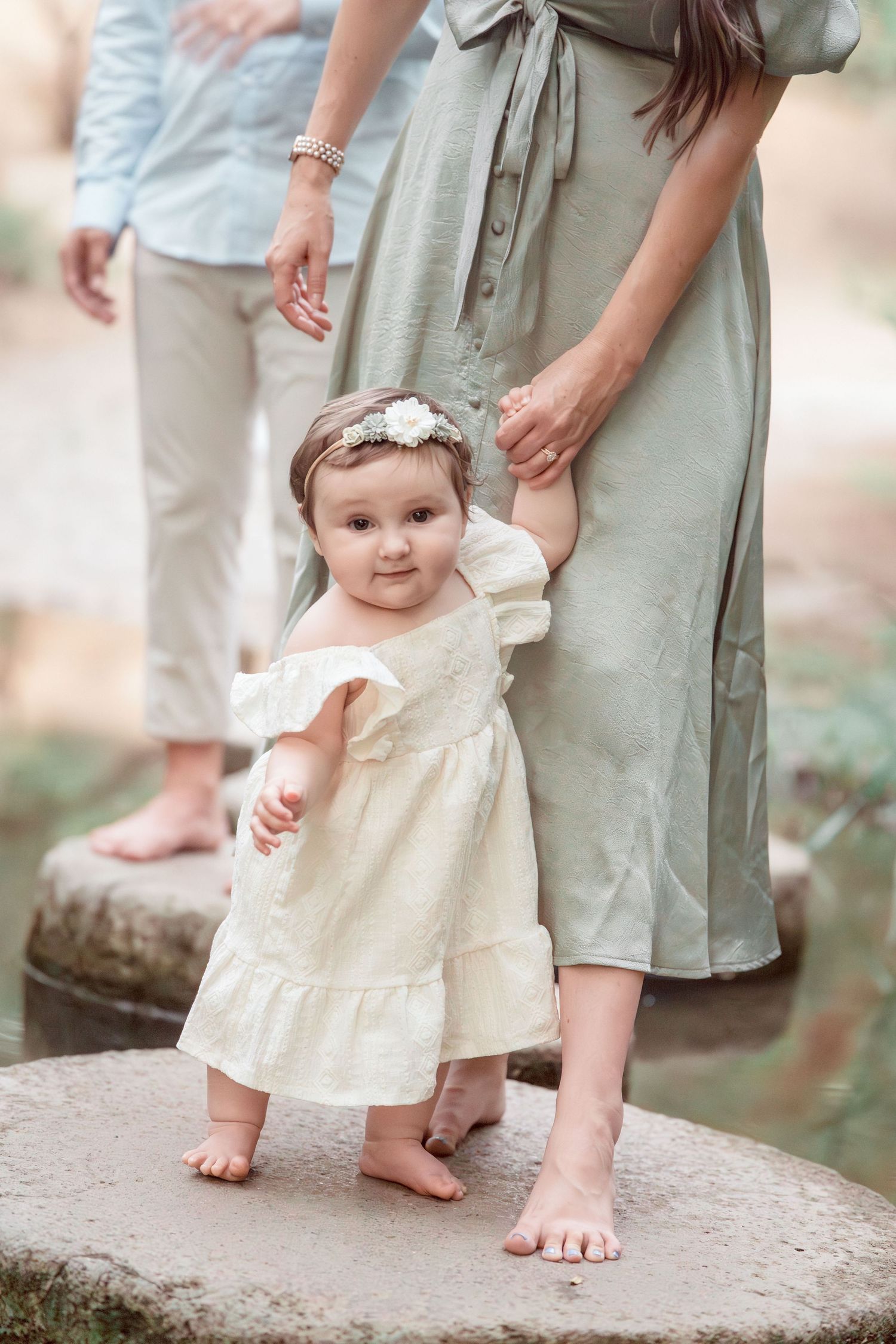 Adorable baby girl in cream dress stands on stepping stone while parents hold her hands in nature setting.