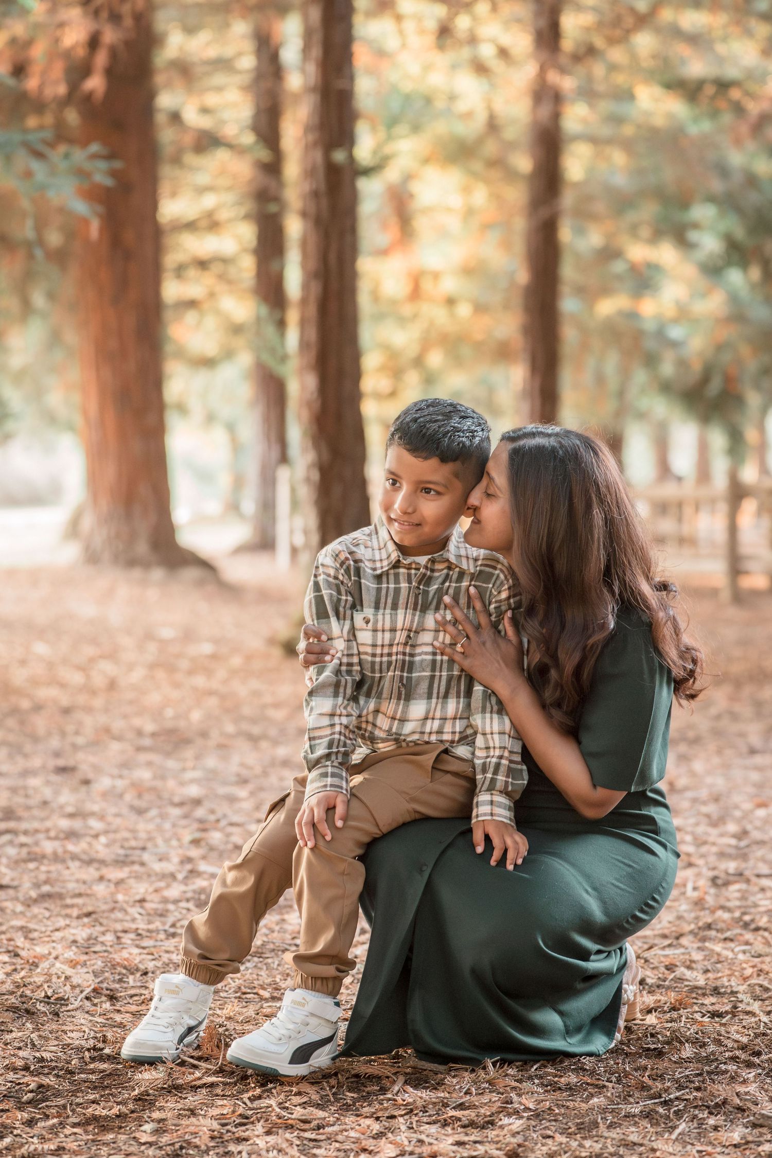 A mother in a green dress sits with her young son on pine needle ground in a warm autumn forest.