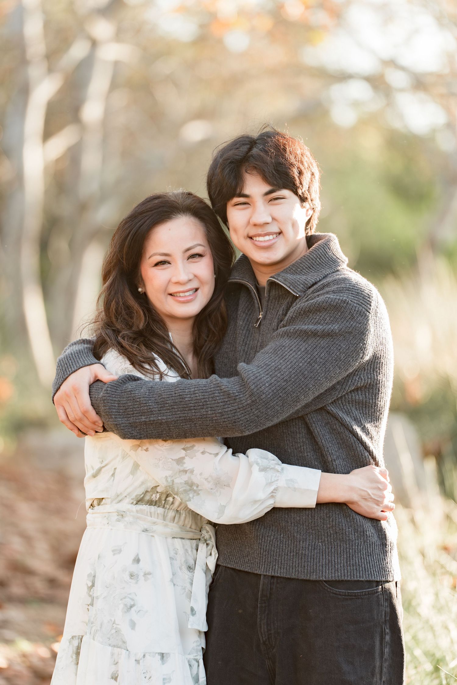 A smiling couple embraces outdoors in a sunlit autumn forest setting, wearing casual attire.