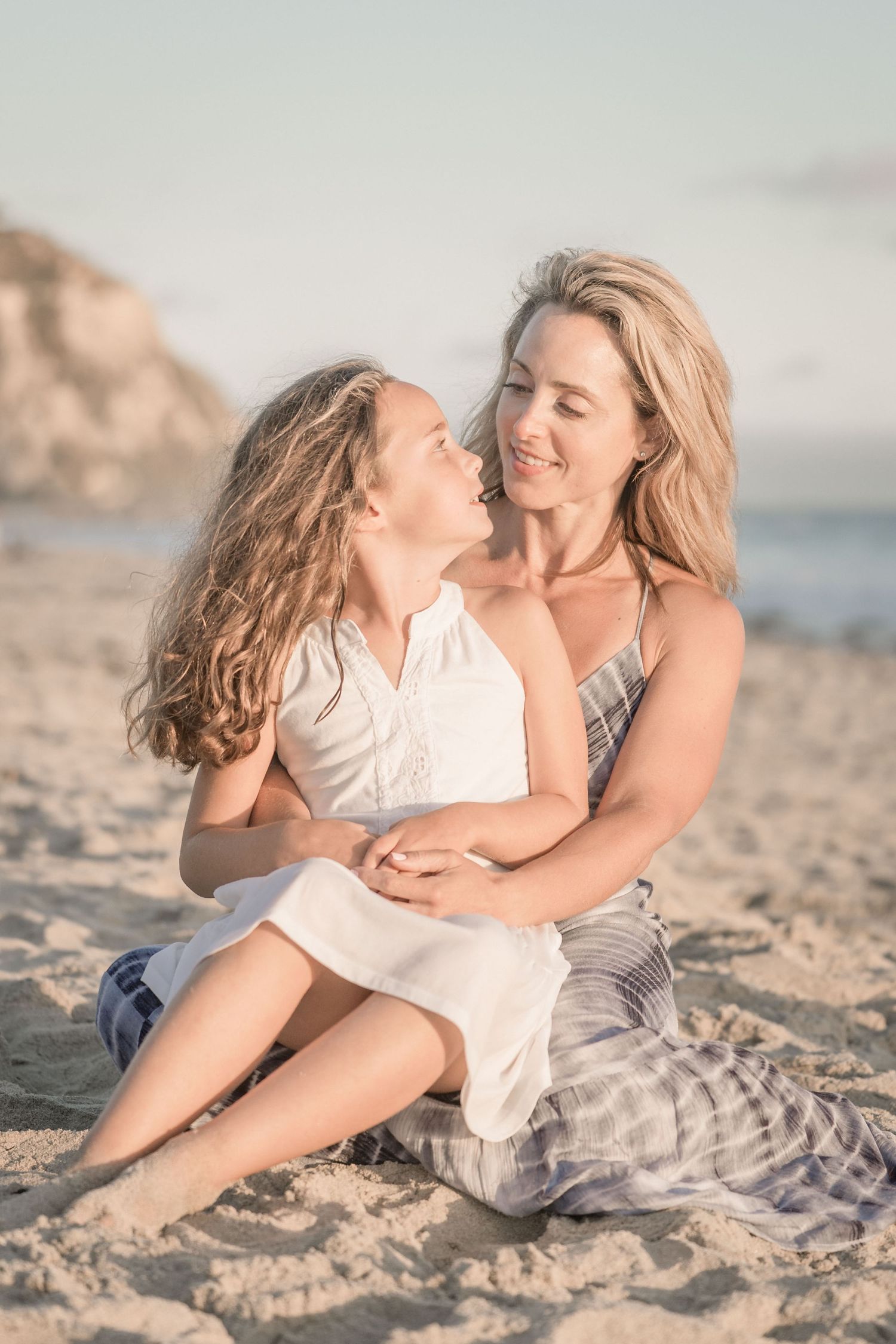 Blonde mother and daughter sharing a tender moment sitting together on sandy beach.