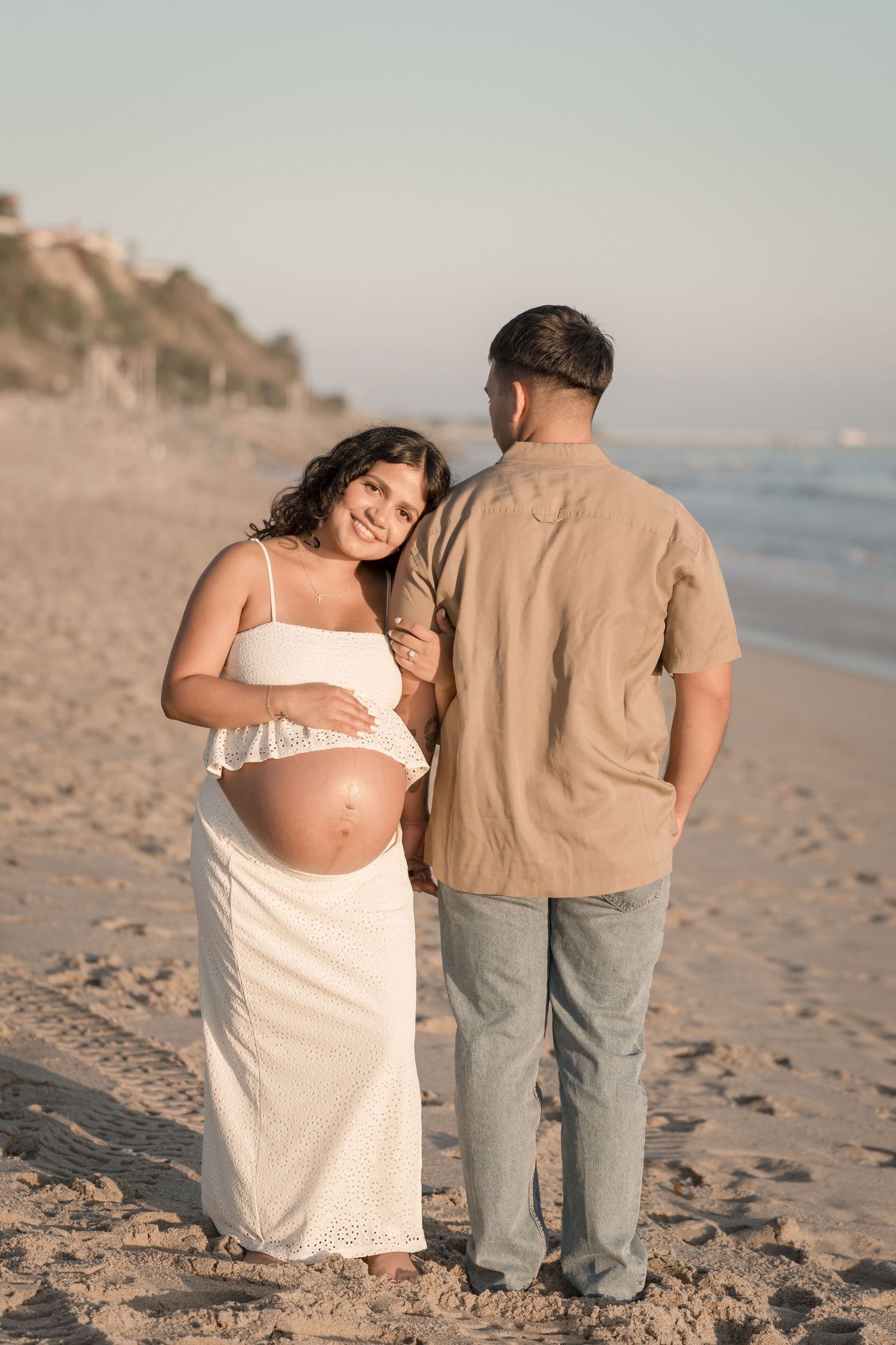 Pregnant woman in white dress with partner on beach at sunset.