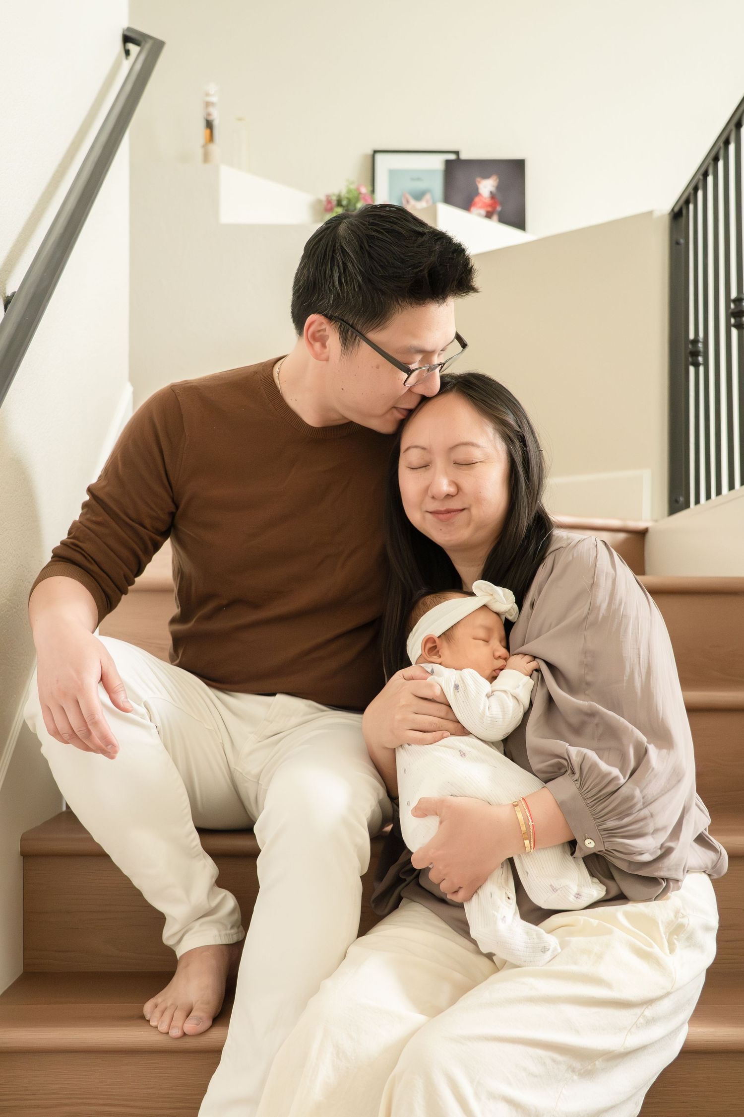Asian couple sitting on home staircase, father kissing mothers head while she holds newborn baby.