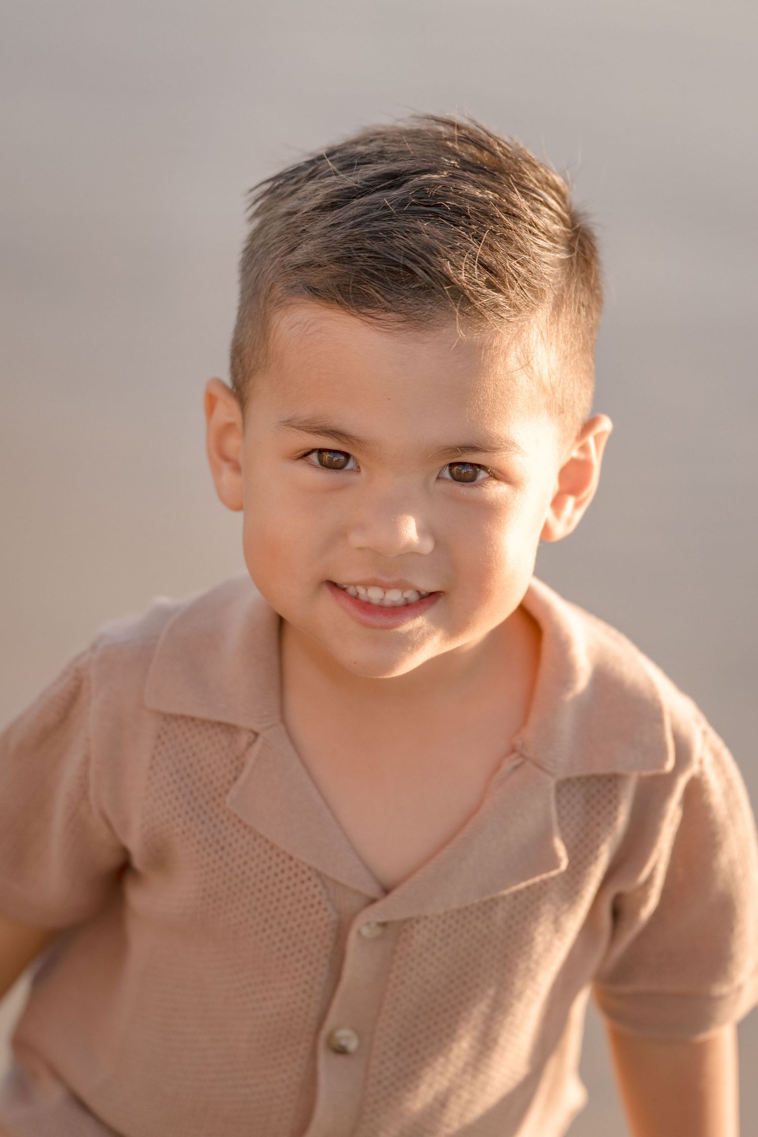 Adorable young boy in a tan polo shirt smiling at the camera during a golden hour beach session.