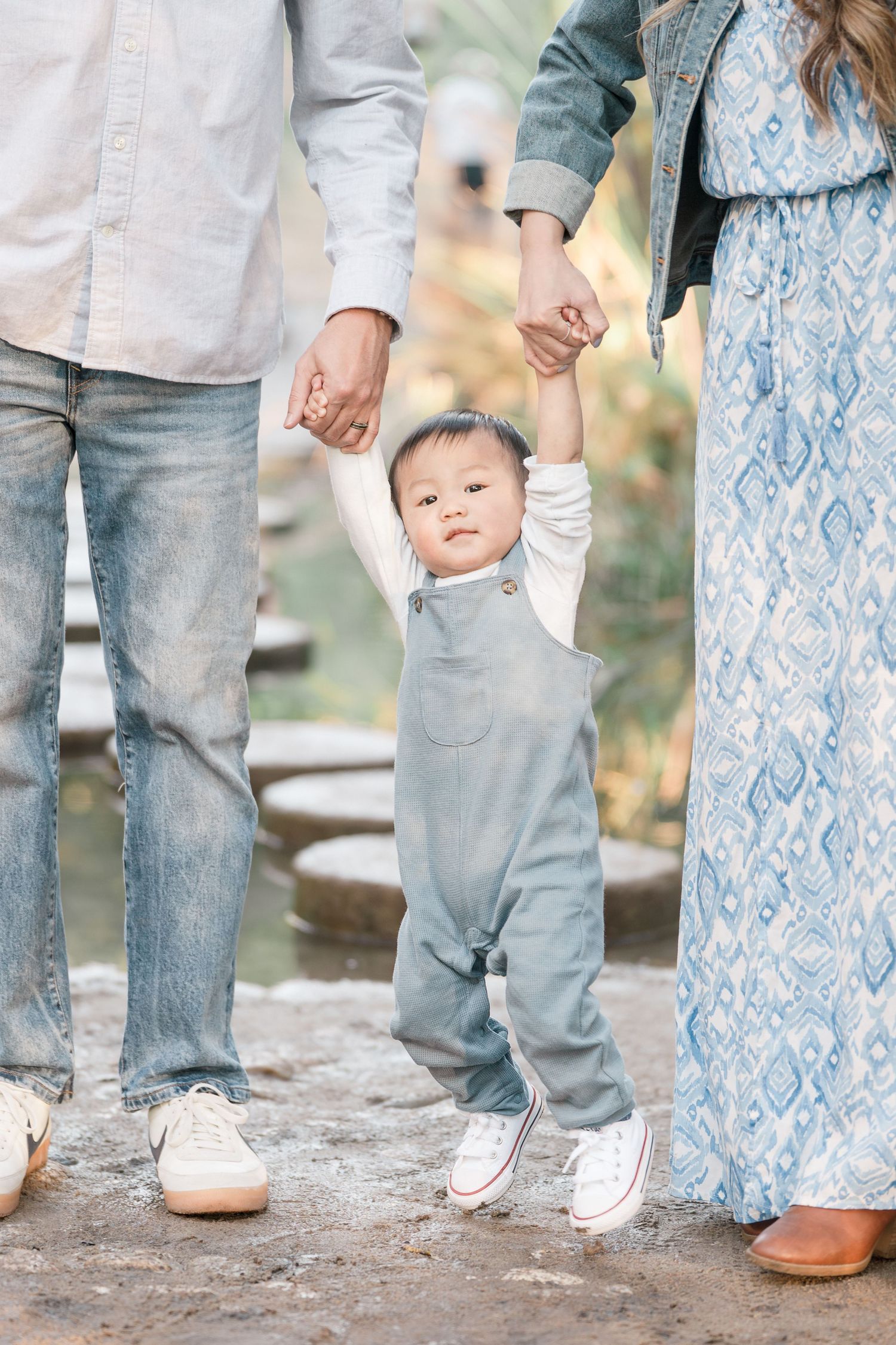 Baby in grey overalls holds parents hands while learning to walk on a garden path.