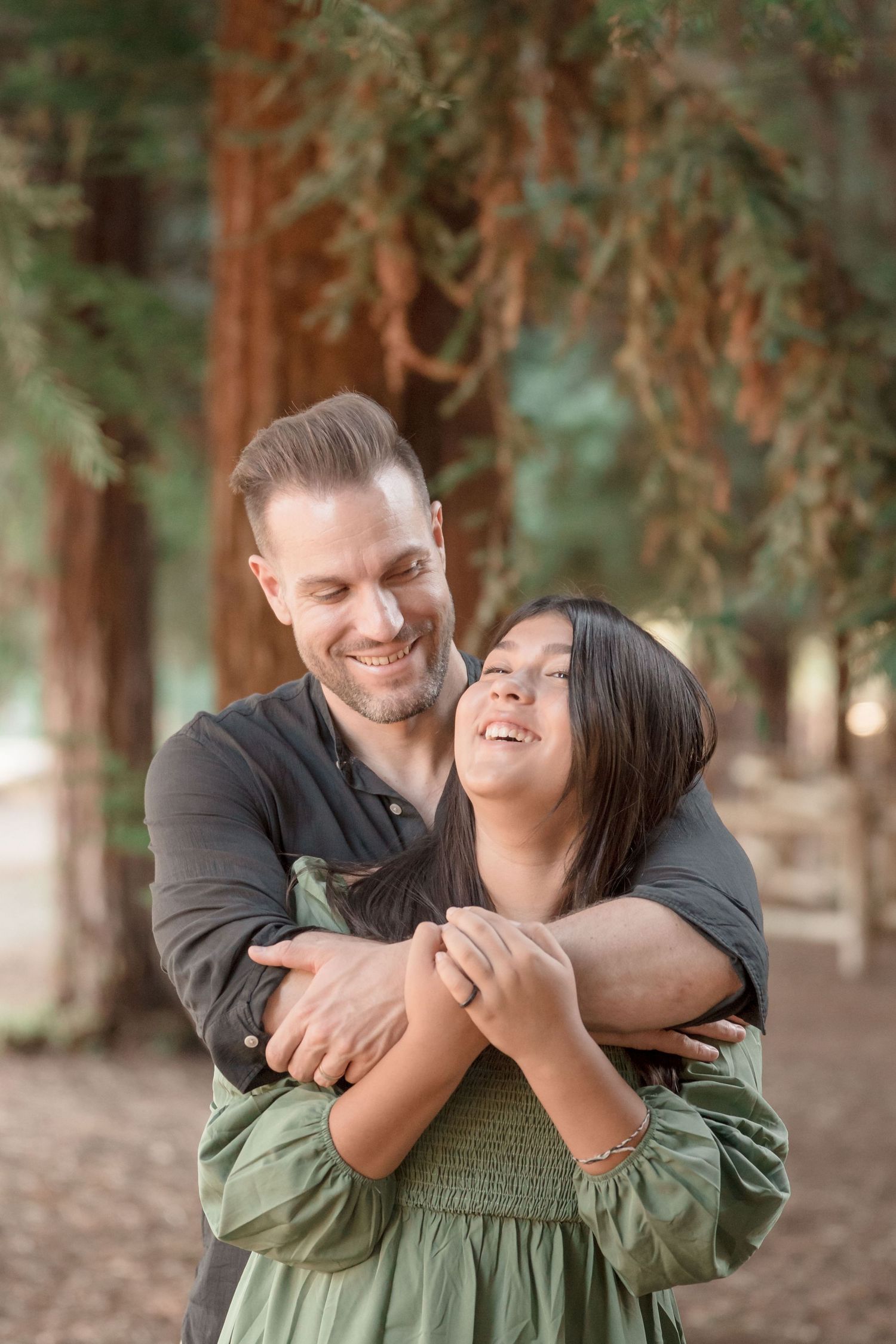 A smiling couple embraces warmly among tall redwood trees in a sunlit forest setting.