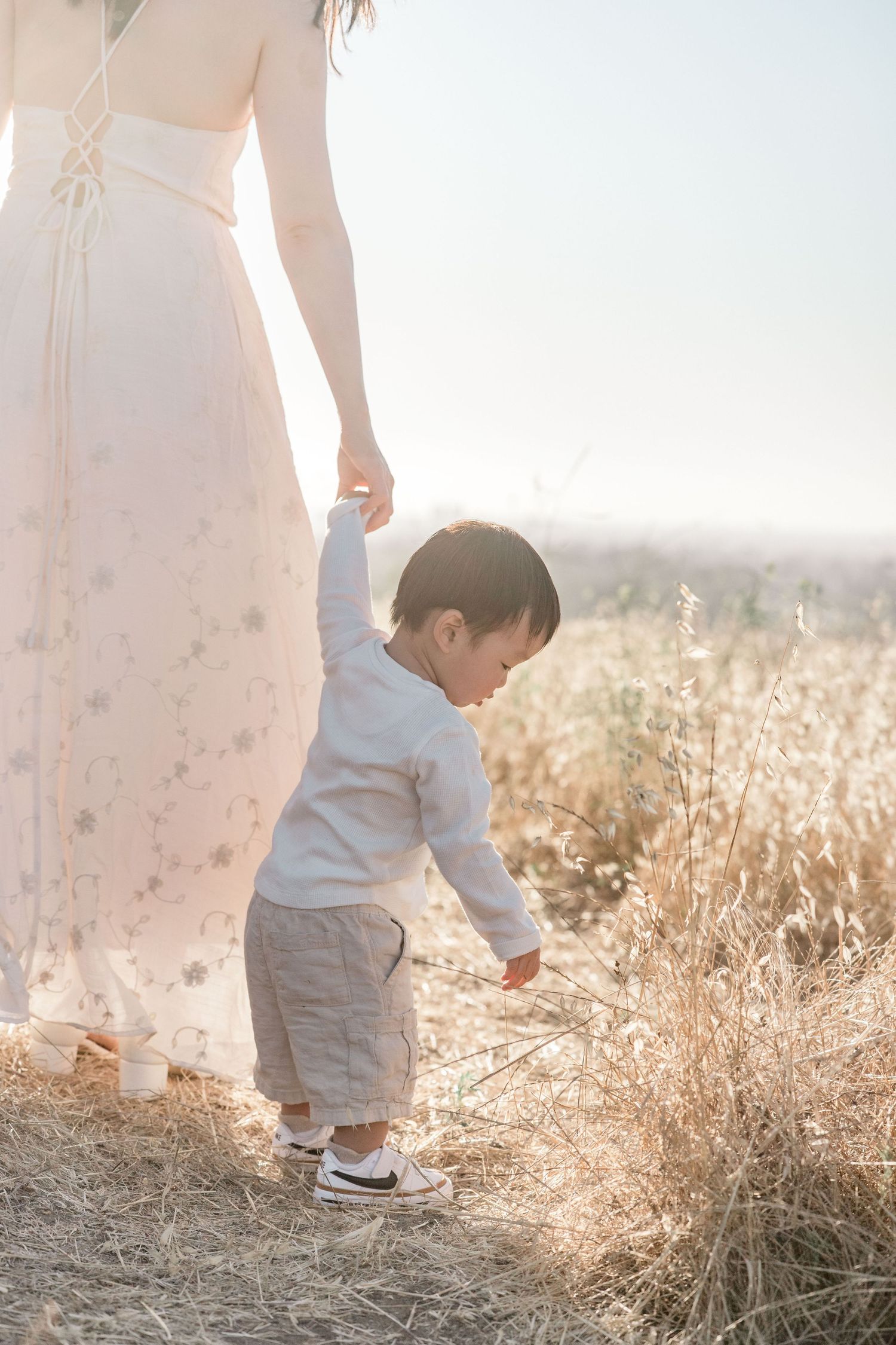 Young toddler holding mothers hand while touching dry grass in a bright sunlit meadow.