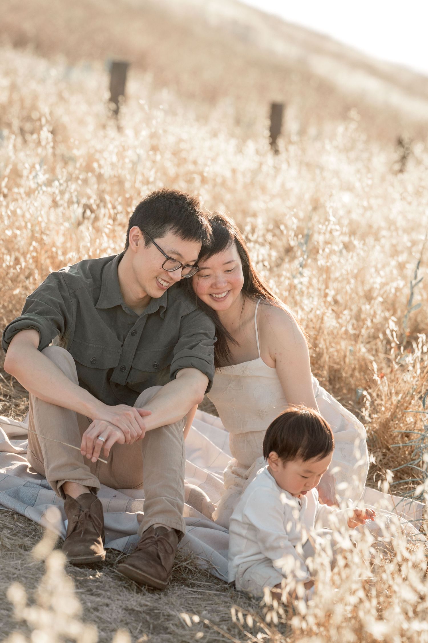Young Asian family relaxing on blanket in golden grass field during warm sunset session.
