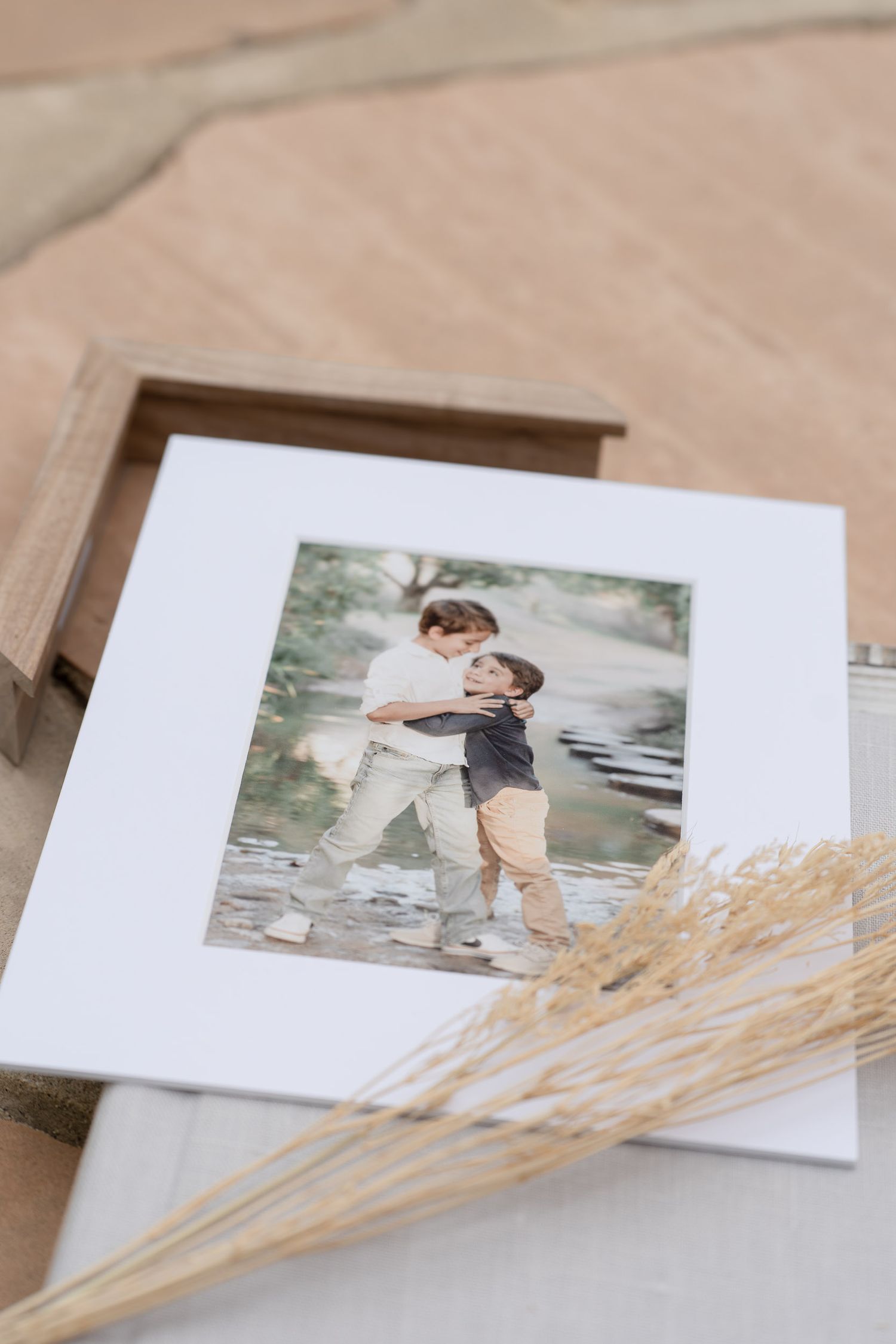A matted photo print of two children playing in water, displayed on a wooden box with dried pampas grass.