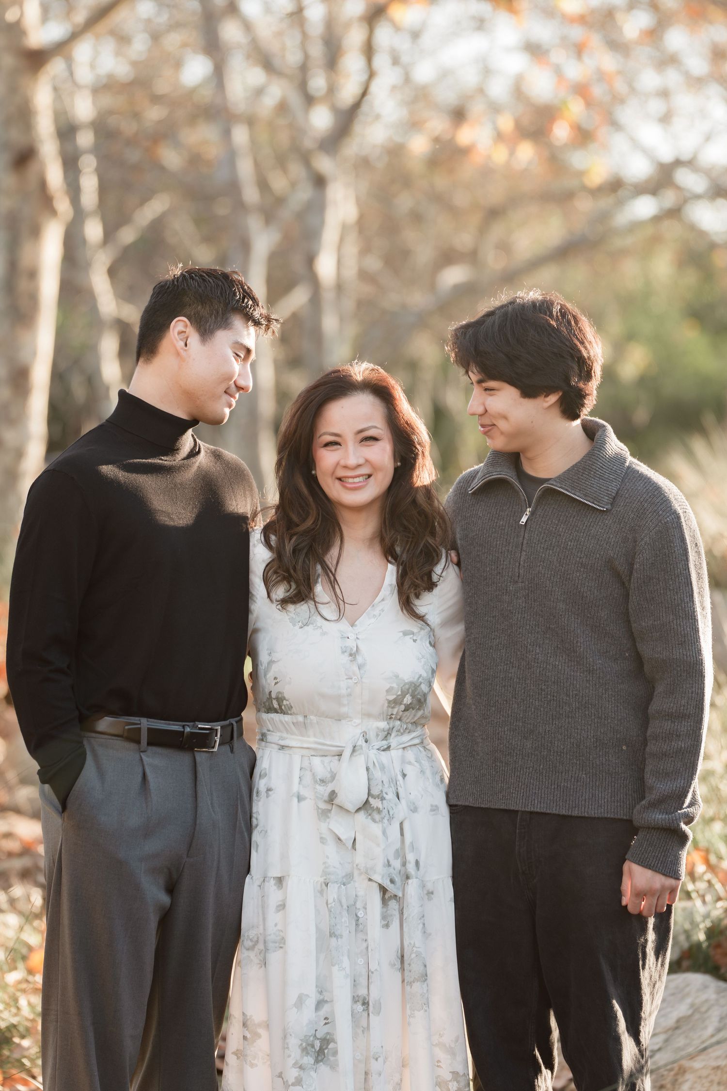 A woman in a floral dress smiles between two young men outdoors in autumn light.