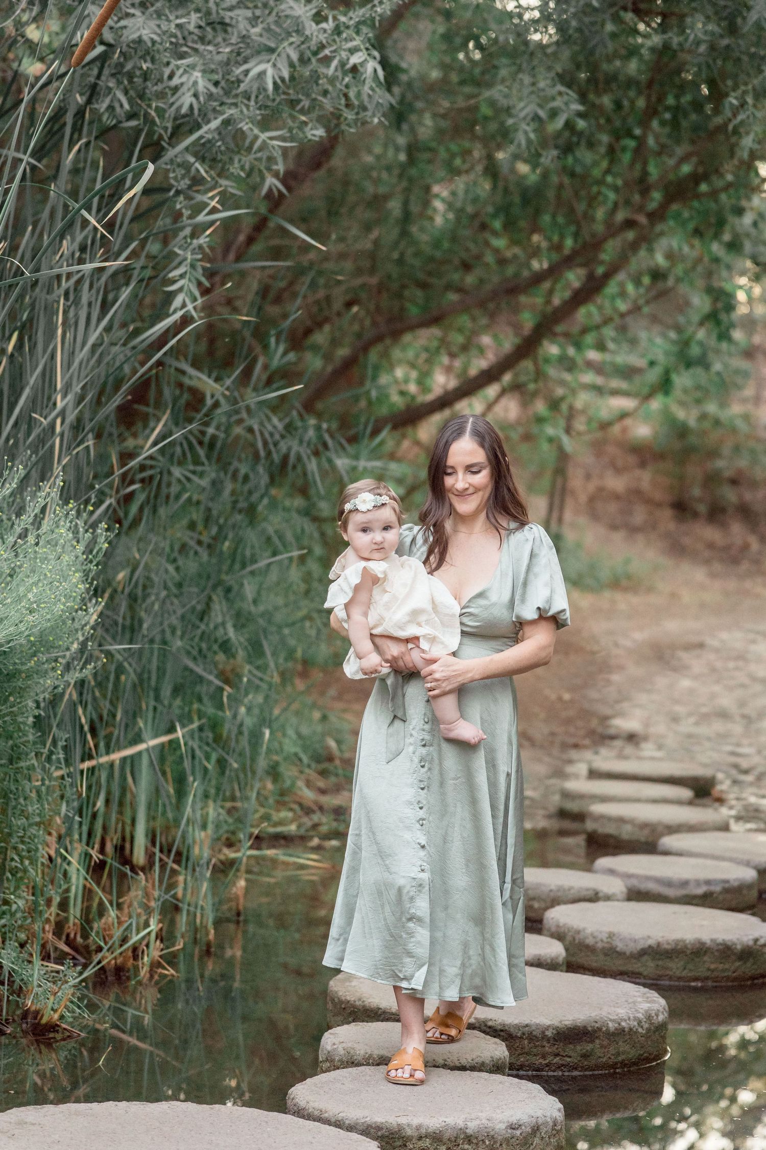 Mother in sage green dress holding baby while standing on stepping stones over a tranquil stream.