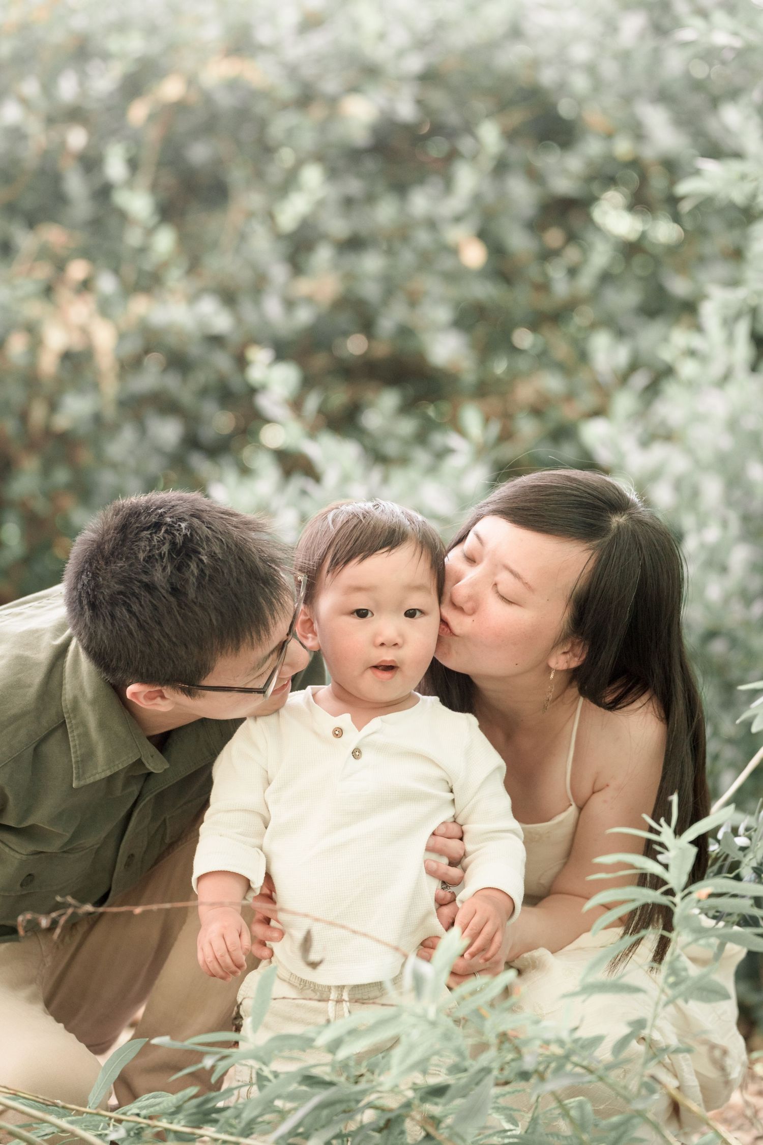 Two parents kissing their baby on the cheeks outdoors, surrounded by lush green foliage in a serene garden setting.