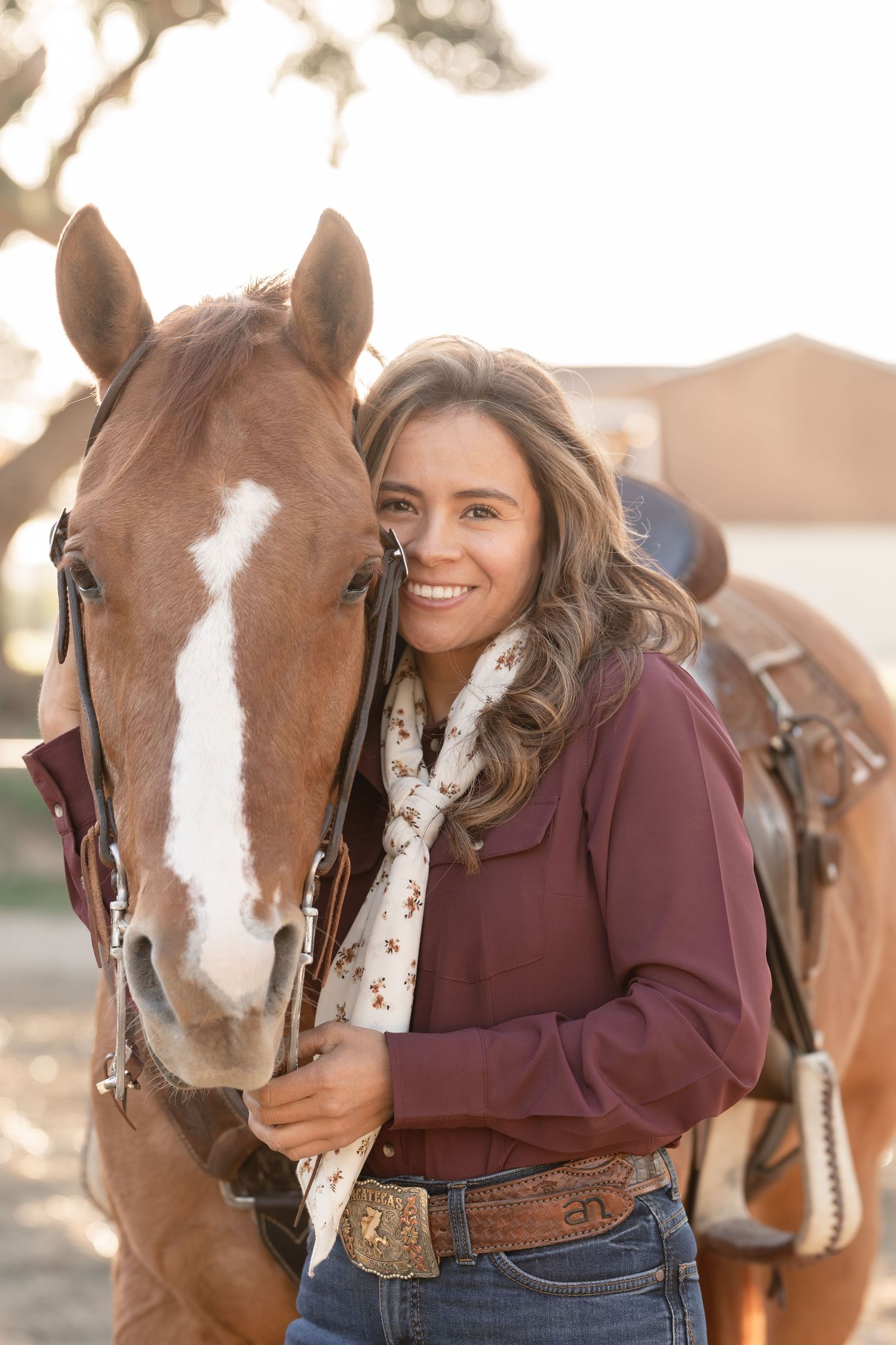 Smiling young woman in western attire hugging a chestnut horse with a blaze marking outdoors.