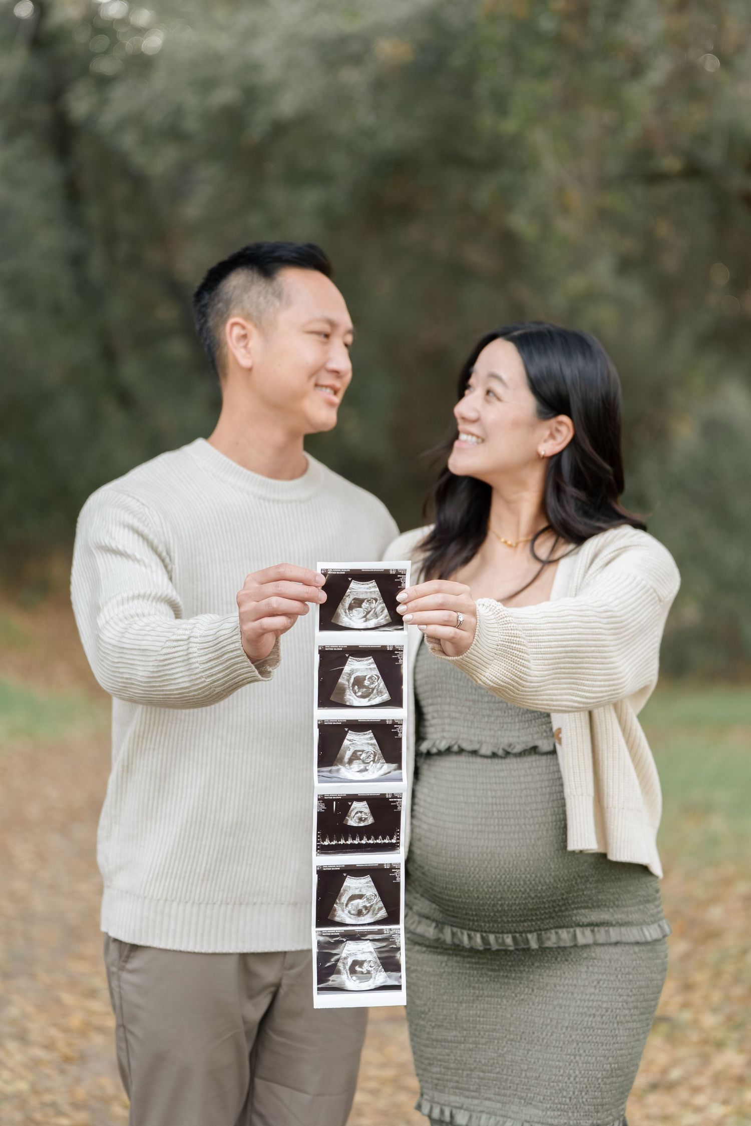 Happy expecting couple holding ultrasound photos outdoors, smiling at each other, woman showing baby bump in green dress.
