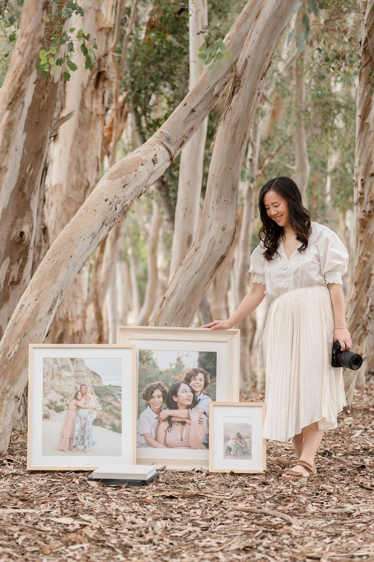 Woman in white dress holding camera poses with framed family portrait prints among eucalyptus trees outdoors.