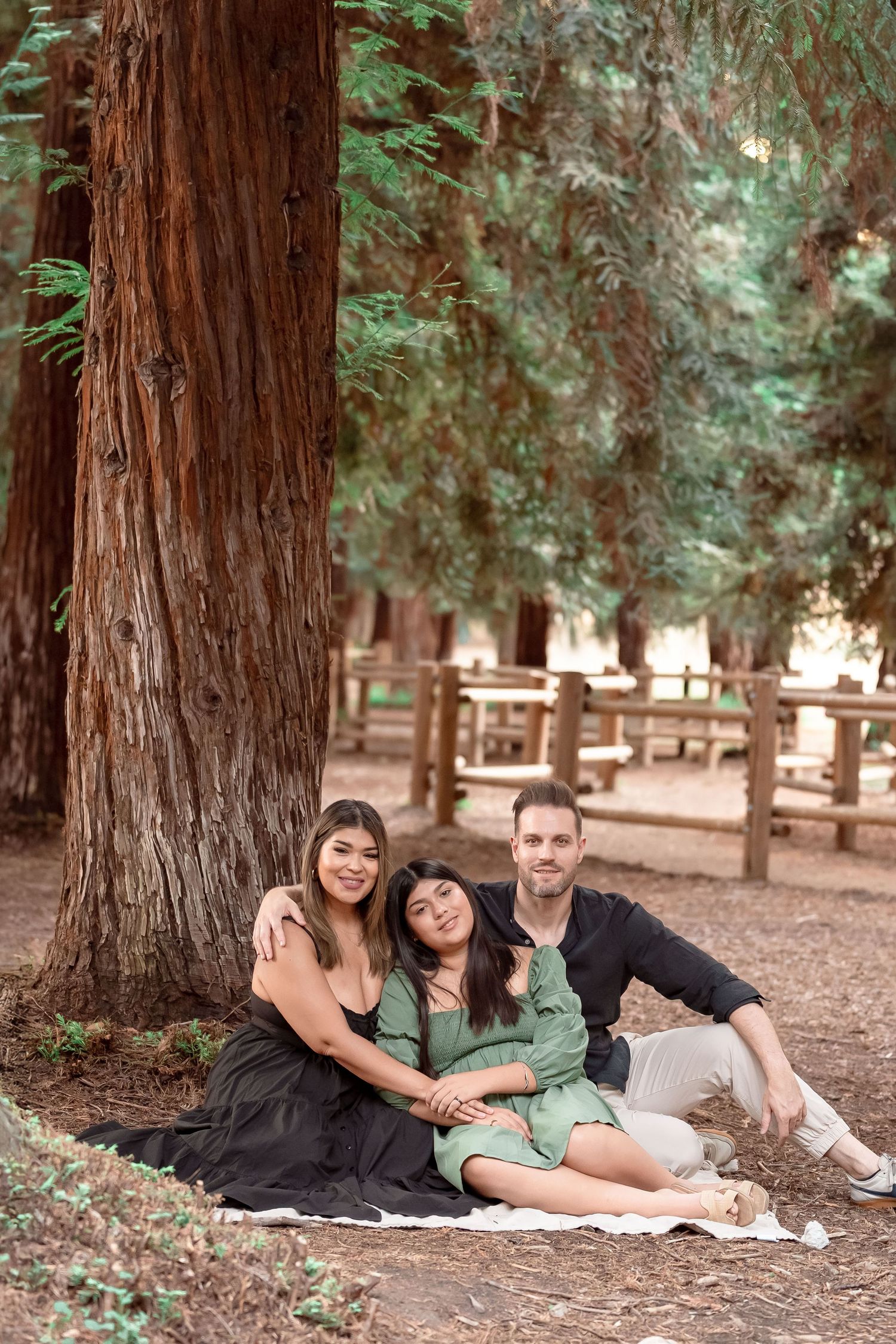 Family of three sitting together on blanket against large redwood tree trunk in forest park.