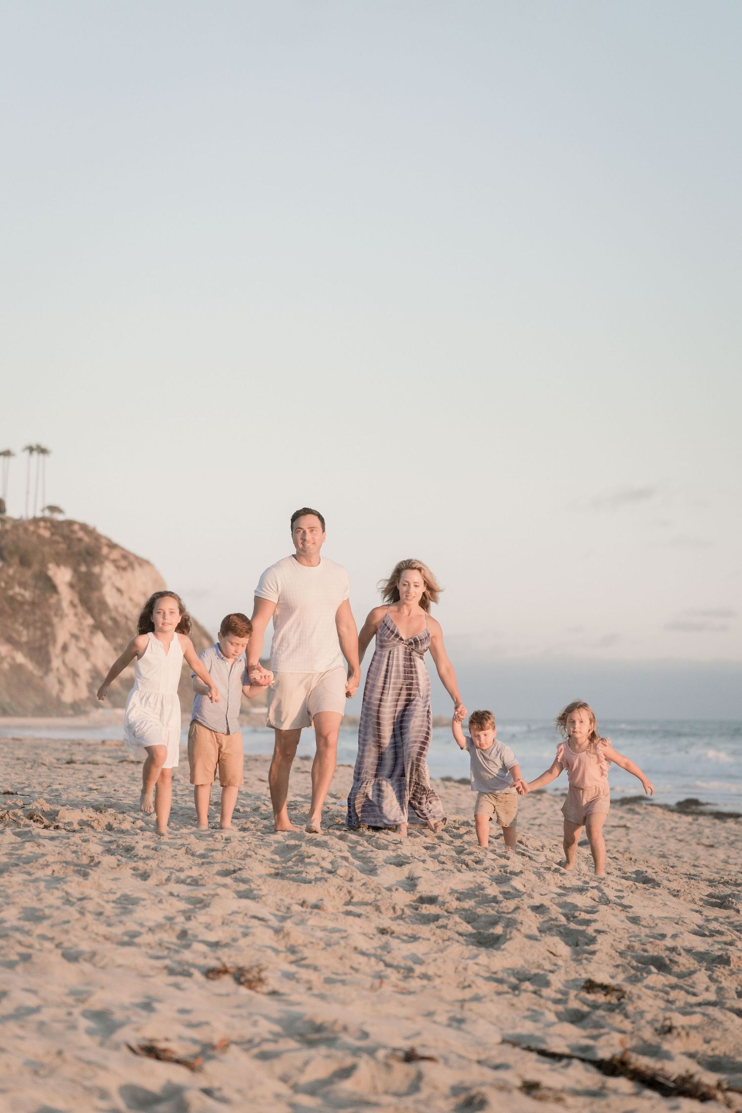 Happy family of six walks hand-in-hand along a sunlit beach at sunset near coastal cliffs.