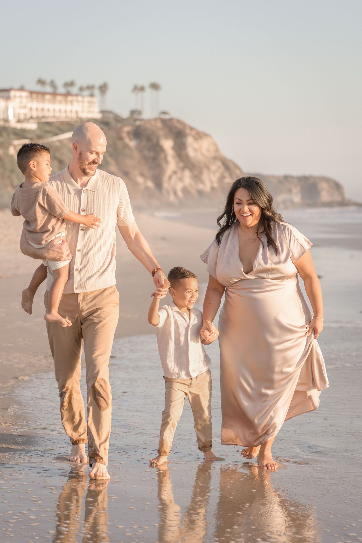 Family of four walking barefoot on a sunny beach, parents holding child's hands, cliffside resort in background.